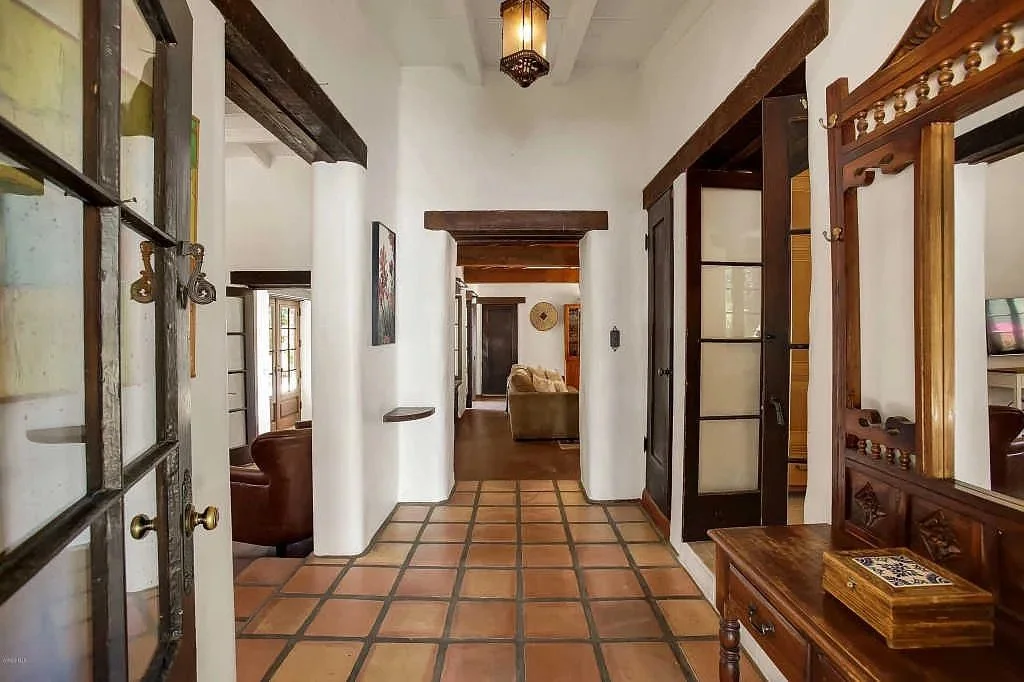 Interior view of a hallway with white walls, wooden beams, terracotta tile floor, and a glimpse into a living room with beige furniture and wooden accents.