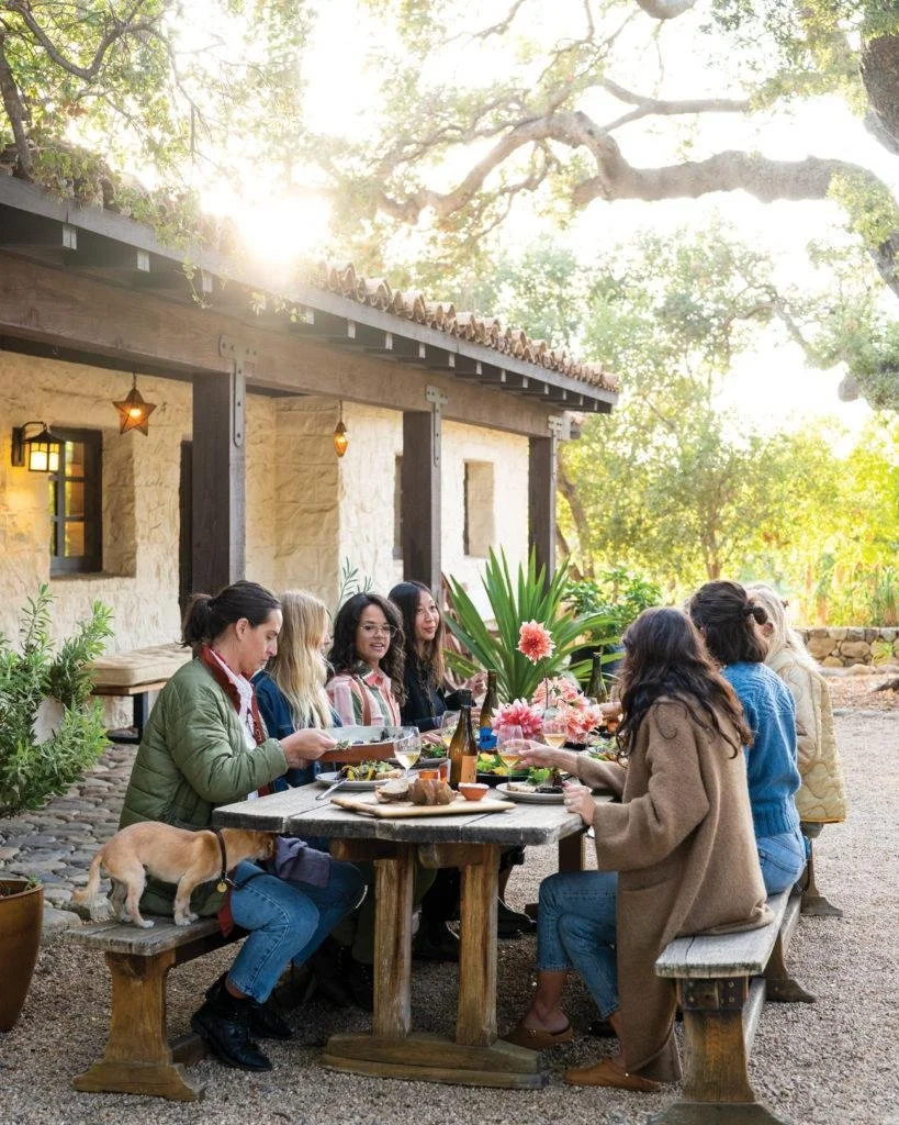 A group of women sitting around an outdoor wooden table, enjoying a meal. The setting appears to be a rustic, outdoor patio surrounded by trees and outdoor lighting, with a building in the background.