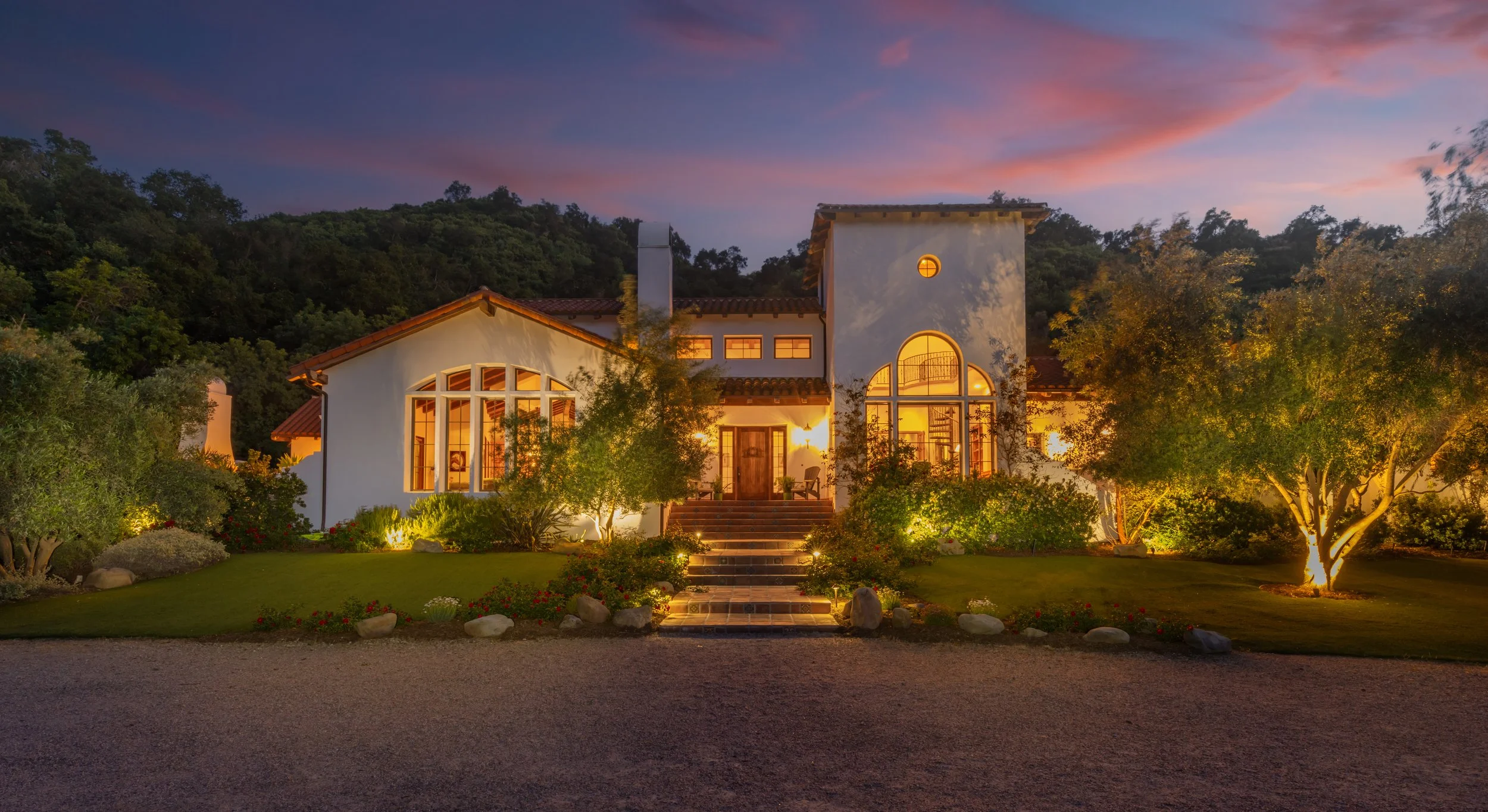 A large white house with a Spanish tile roof, large arched windows, and a front porch, illuminated at dusk with the sky showing pink and purple hues. The house is surrounded by trees and a well-maintained garden with landscape lighting.