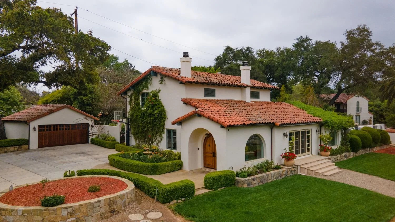 A large Mediterranean-style house with white stucco walls, red tile roof, arched doorway, and multiple chimneys. The house is surrounded by well-manicured lawn, shrubs, and potted plants. There is a driveway and a detached garage with a wooden door. Trees and neighboring houses are visible in the background.