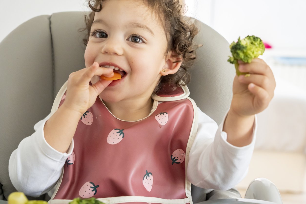 A young girl with curly hair eating a carrot and holding a piece of broccoli while sitting in a highchair, wearing a strawberry-patterned bib, working on chewing and swallowing skills during feeding therapy for kids in Lemont, IL.