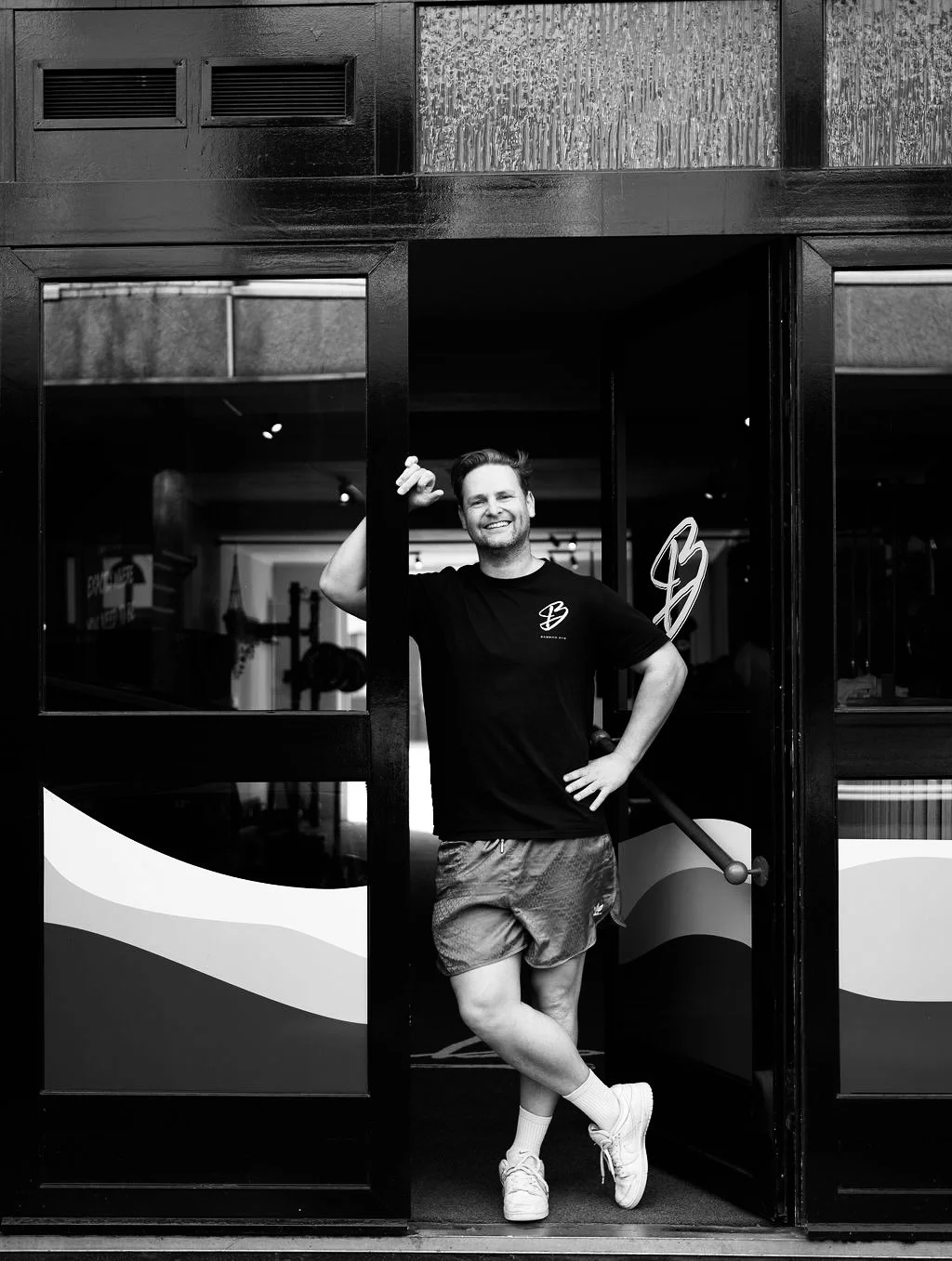 Man staat in de deuropening, lachend, met één hand in de zij en de andere hand omhoog. Draagt een zwart T-shirt en korte broek, binnen een fitnessruimte.