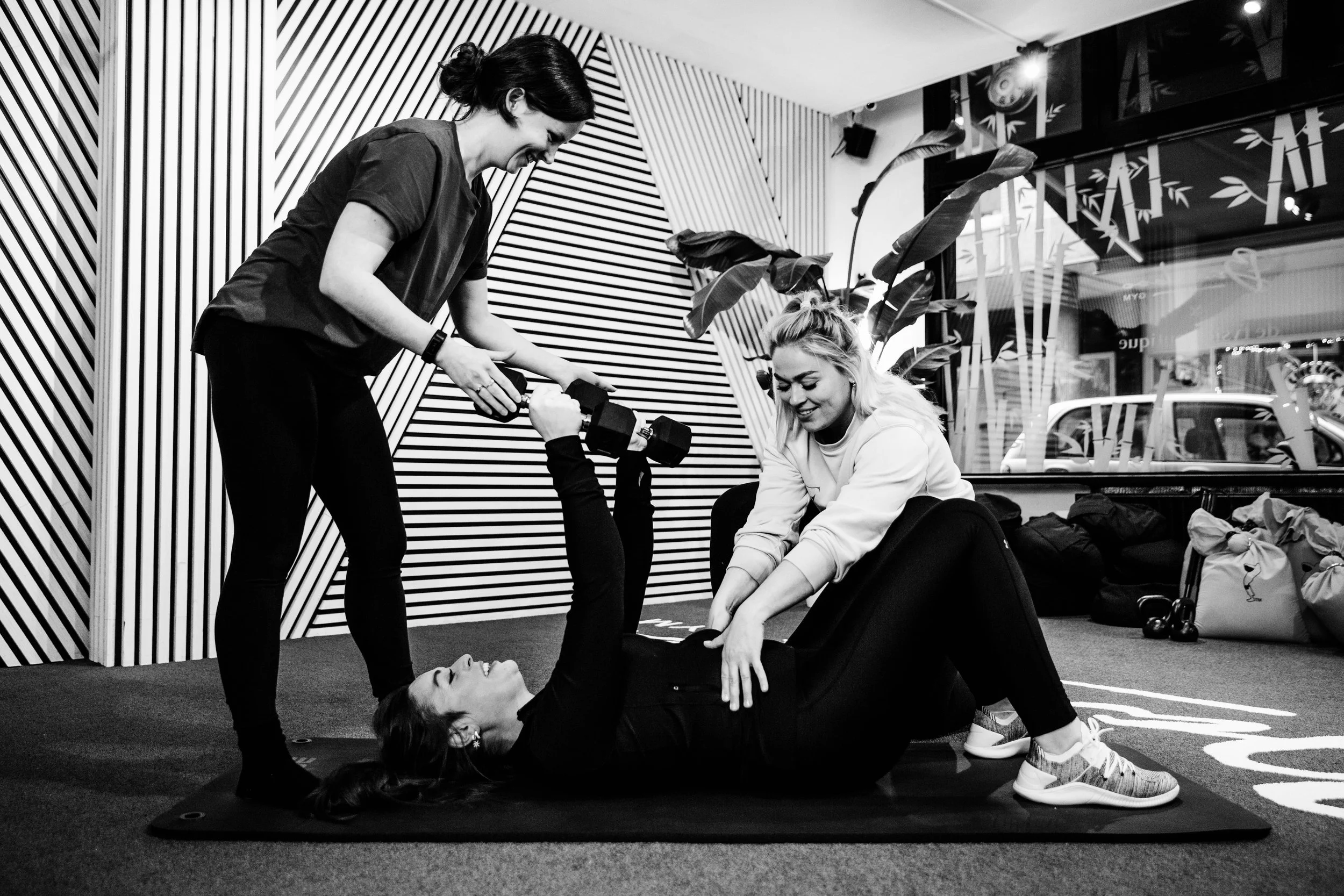Vrouw ligt op de grond terwijl twee andere vrouwen haar helpen tijdens een yogales in een studio met decoratieve wand en grote ramen.
