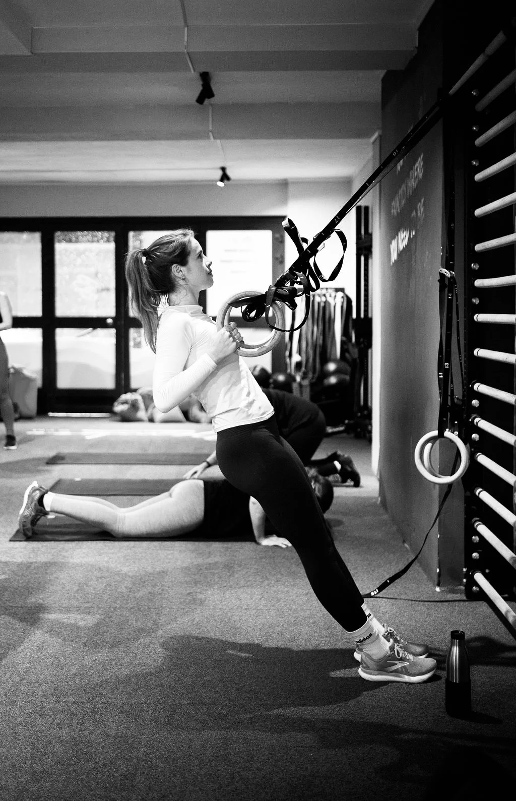 Een vrouw doet oefeningen met gimnastrings in een sportschool, omgeven door andere mensen die ook sporten.
