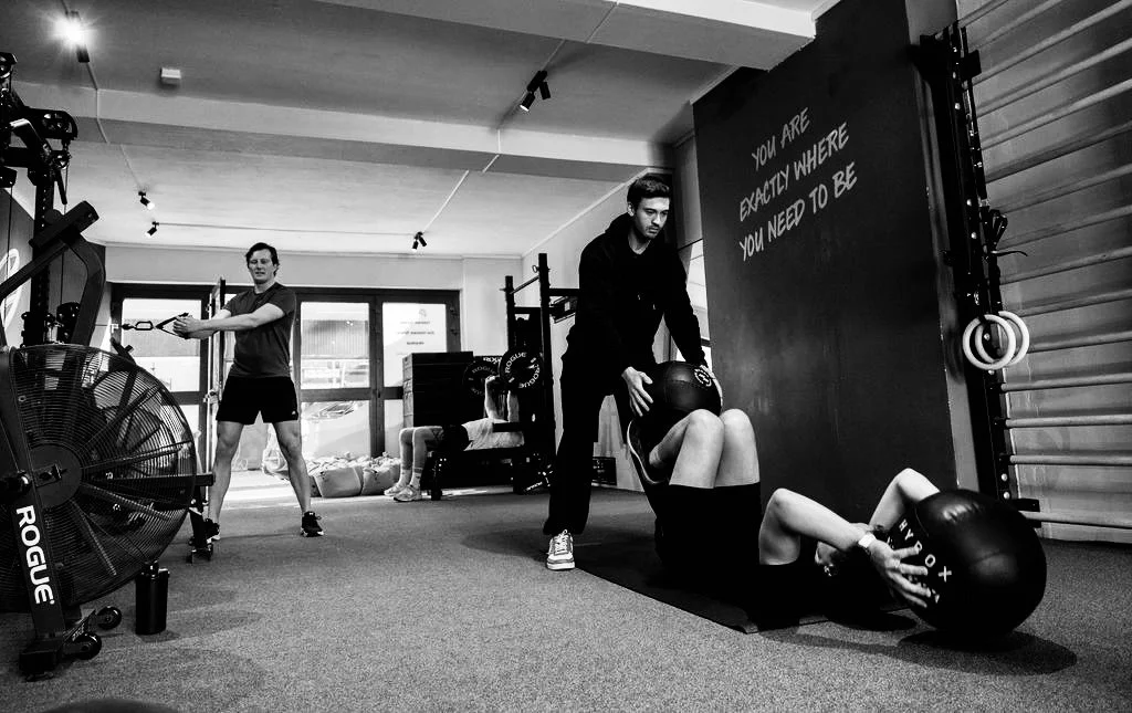 Twee personen die in een sportschool trainen met medicijnballen, een persoon ligt op de grond terwijl de andere helpt, een derde persoon traint op de achtergrond.