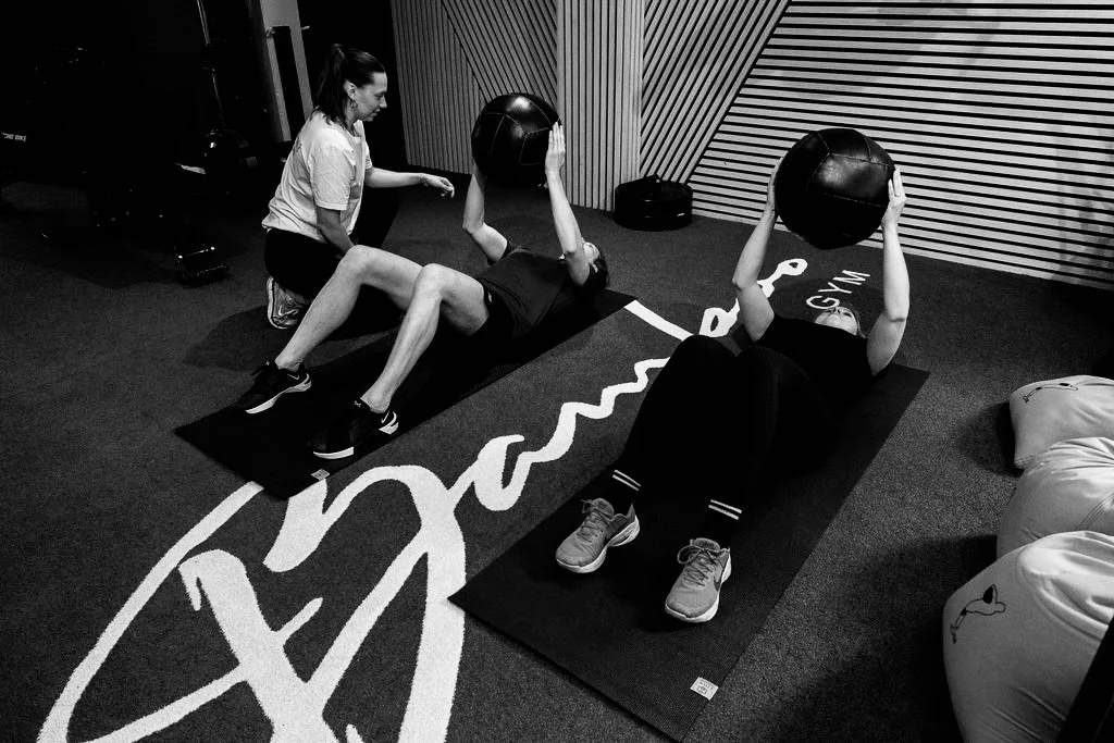 Twee mensen doen sit-ups met medicijnballen in een sportschool, terwijl een trainer toekijkt.