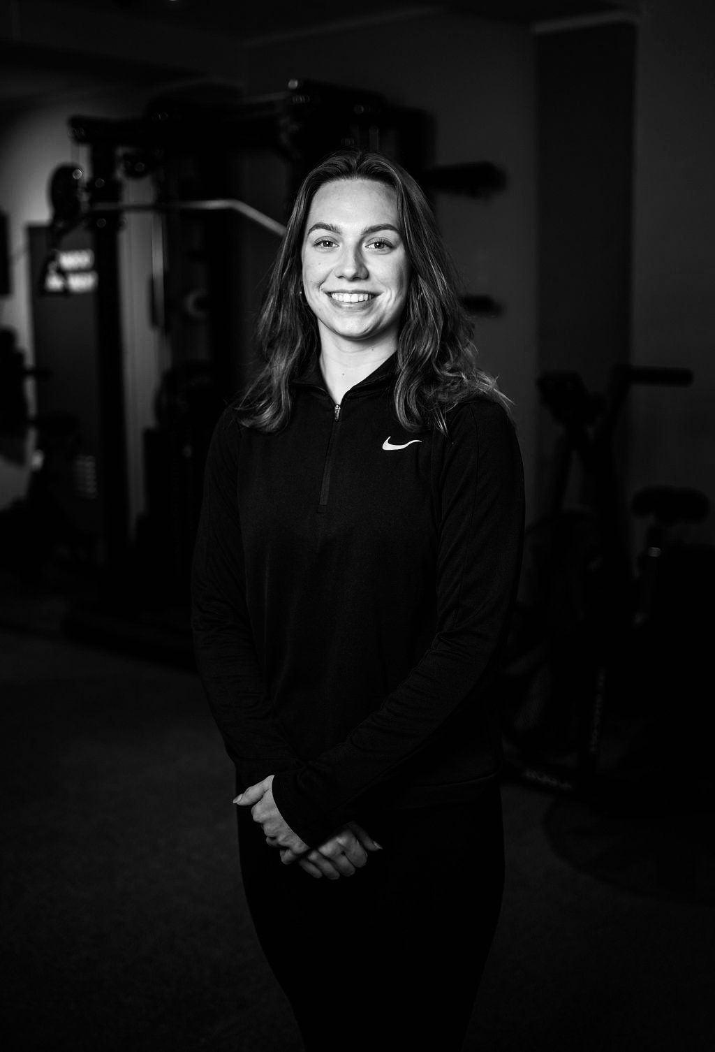 Zwart-wit foto van een lachende vrouw in sportkleding, binnen een fitnessruimte.