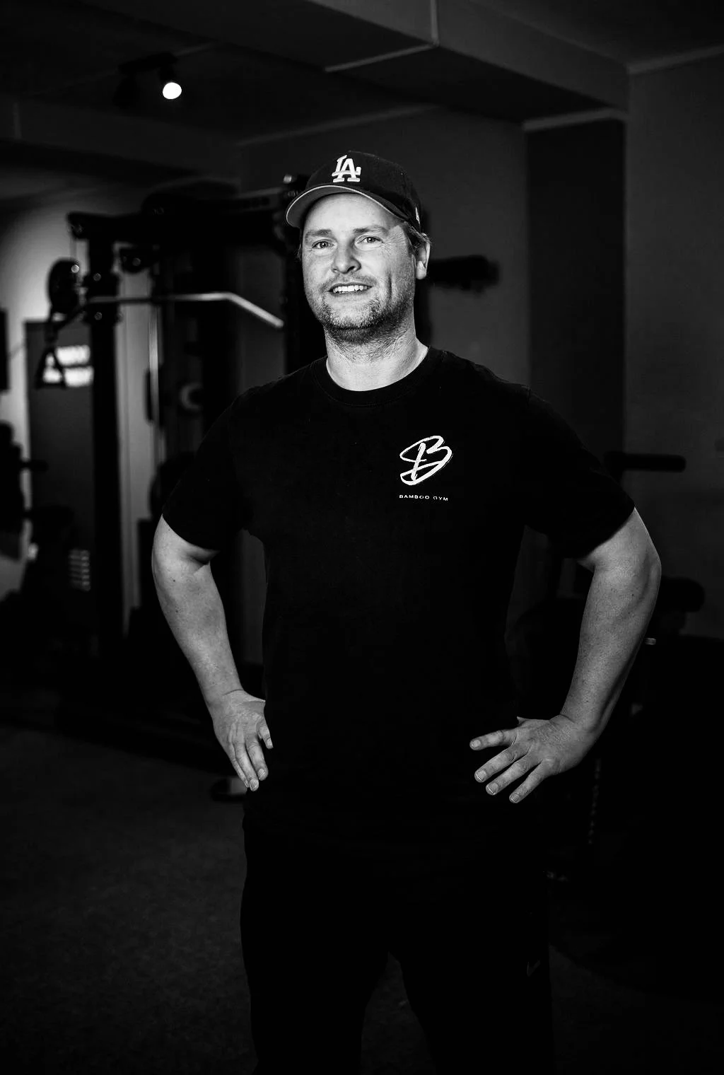 Man in een sportschool, zwart-wit foto, draagt een zwarte T-shirt met een logo en een zwart pet met 'LA' erop.
