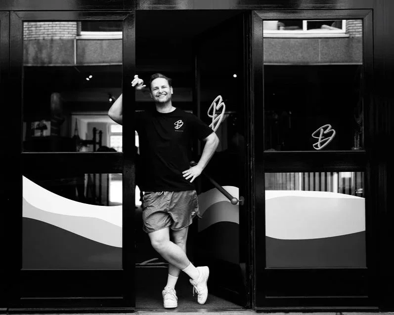 Man staat ontspannen in de deuropening van een café, gekleed in sportkleding, glimlachend met hand in haar haar en arm op de heup, zwart-witfoto.