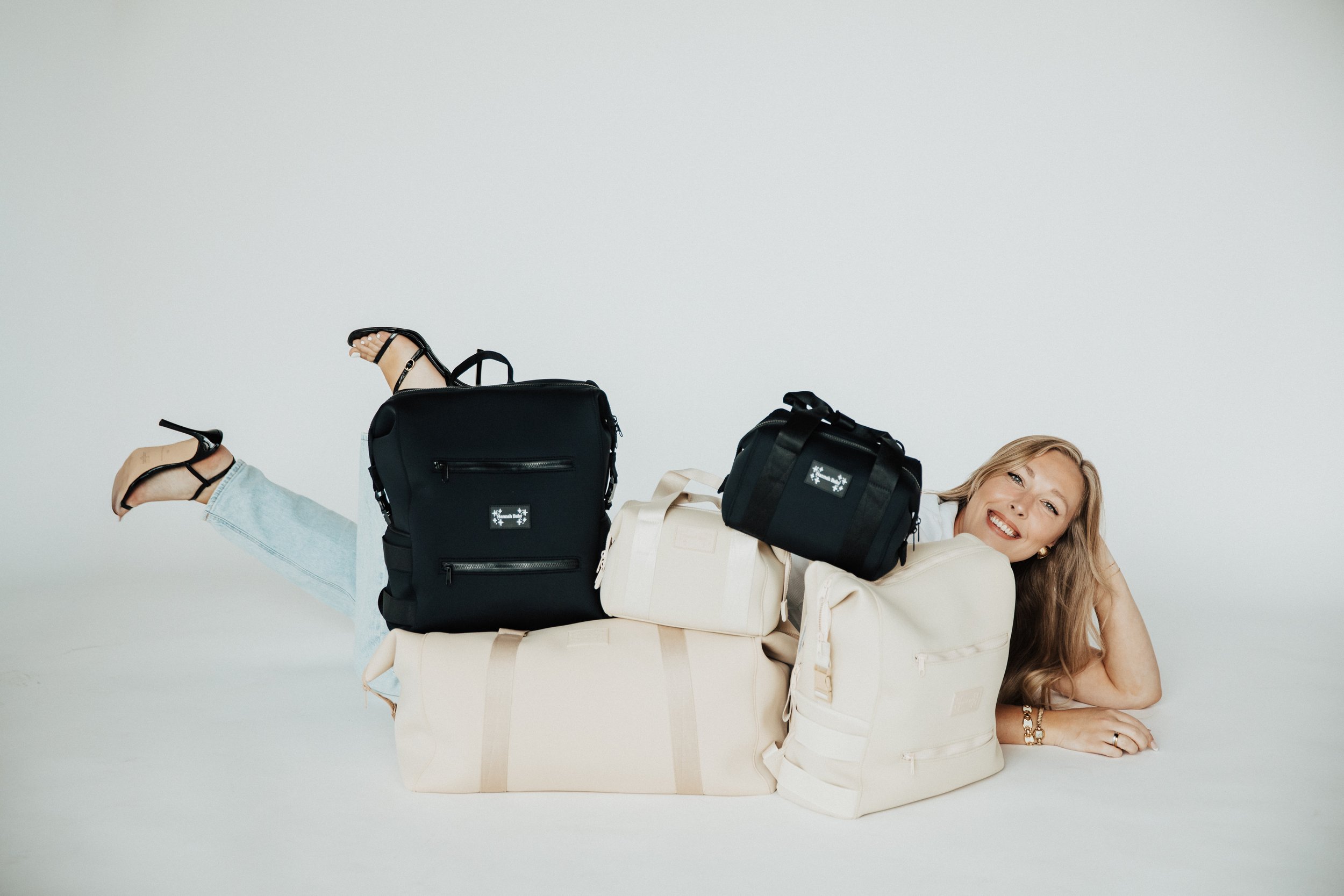 A young woman with long wavy hair lying behind a stack of beige and black luggage, smiling at the camera, with multiple black and beige diaper and pump bags placed on and around her, against a plain white background.