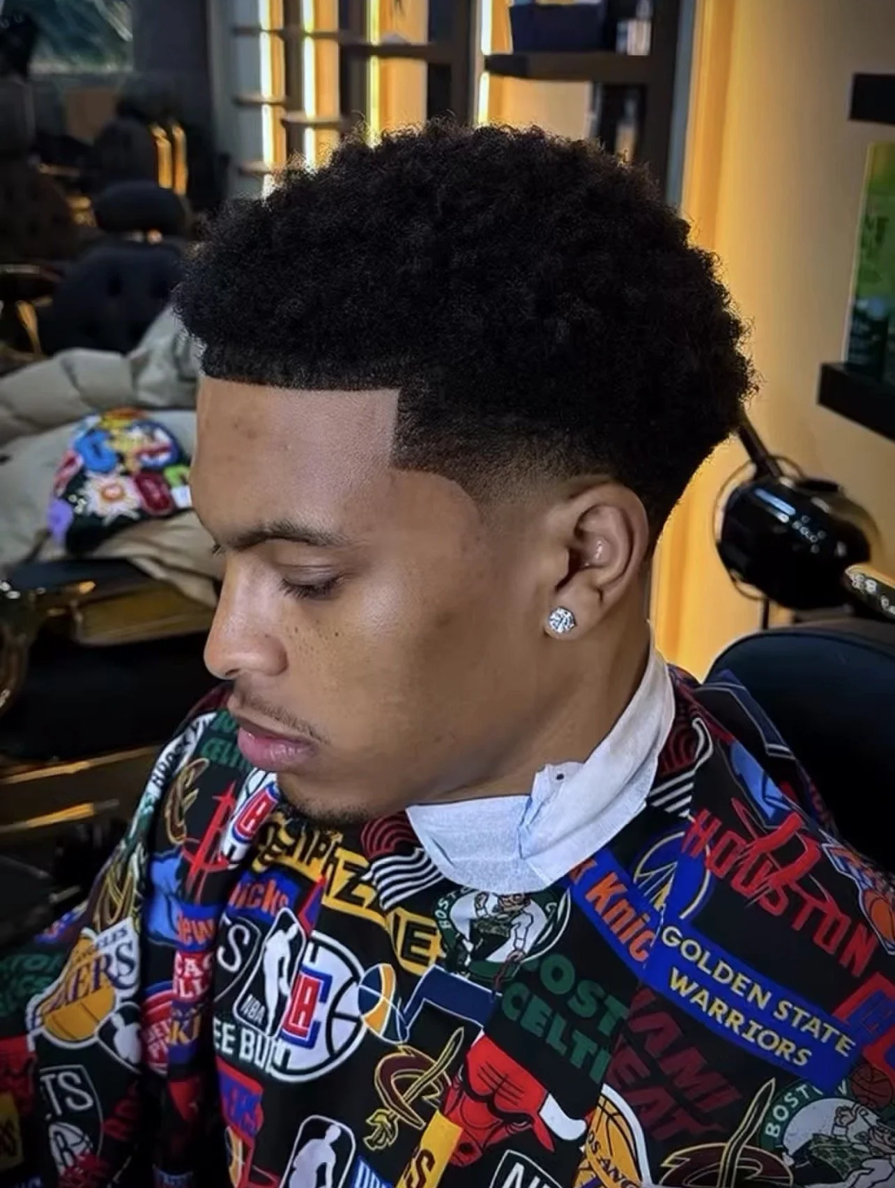Young man with curly hair and diamond earring in a barbershop, wearing a colorful sports-themed cape and a face mask hanging below his chin.
