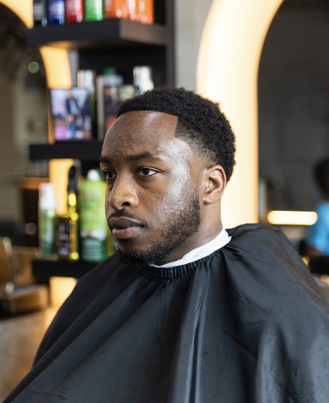 A young man with a fade haircut and beard sitting in a barbershop chair, wearing a black cape, with colorful hair products on shelves in the background.