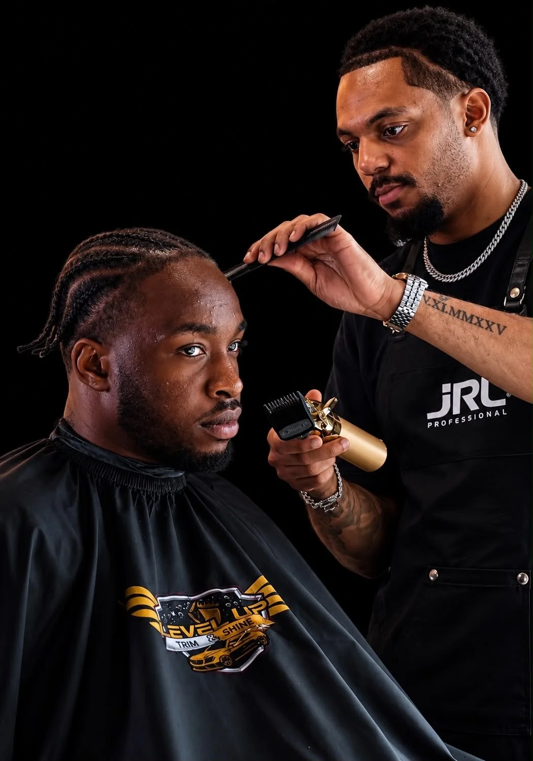 A Black man sitting in a barber's chair getting a haircut, with braided hair, while a barber trims his hair with professional clippers. The barber is focused on his work, wearing a black apron with Donnelley's logo, and has a tattooed arm, a watch, and chain necklaces.