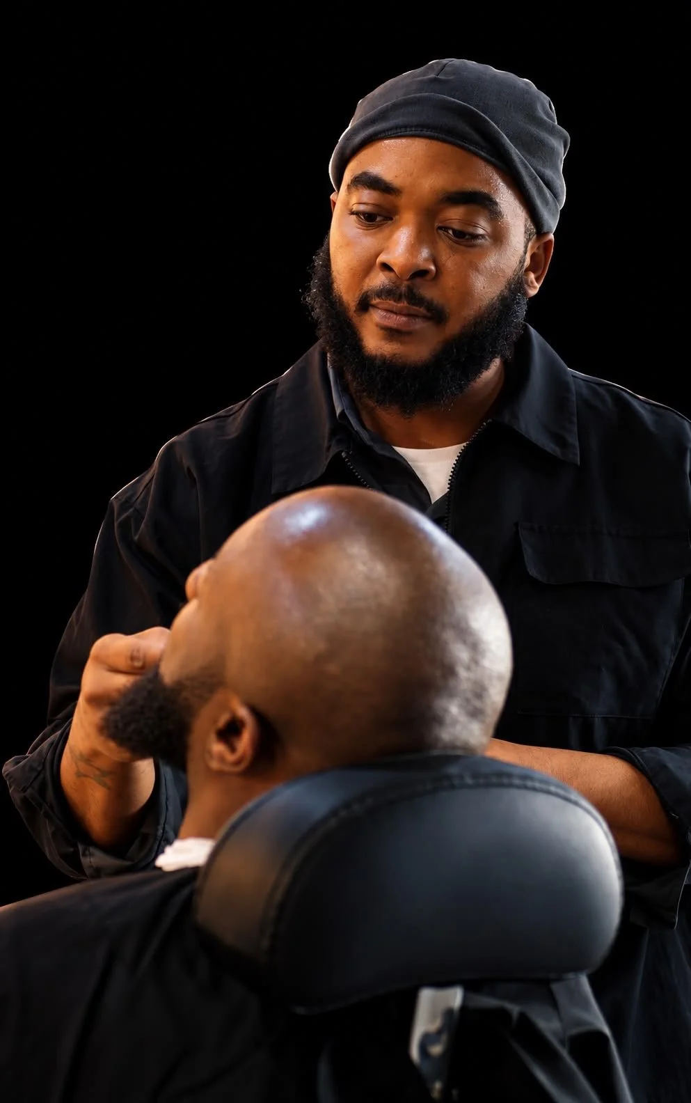 A black man with a beard wearing a gray beanie and black jacket is giving a haircut to a client with a bald head, black chair, and black background.