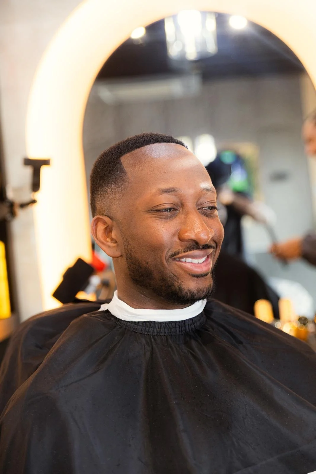 Man in barber shop sitting in chair, smiling with freshly cut hair and beard, wearing a black barber cape, mirror in the background.