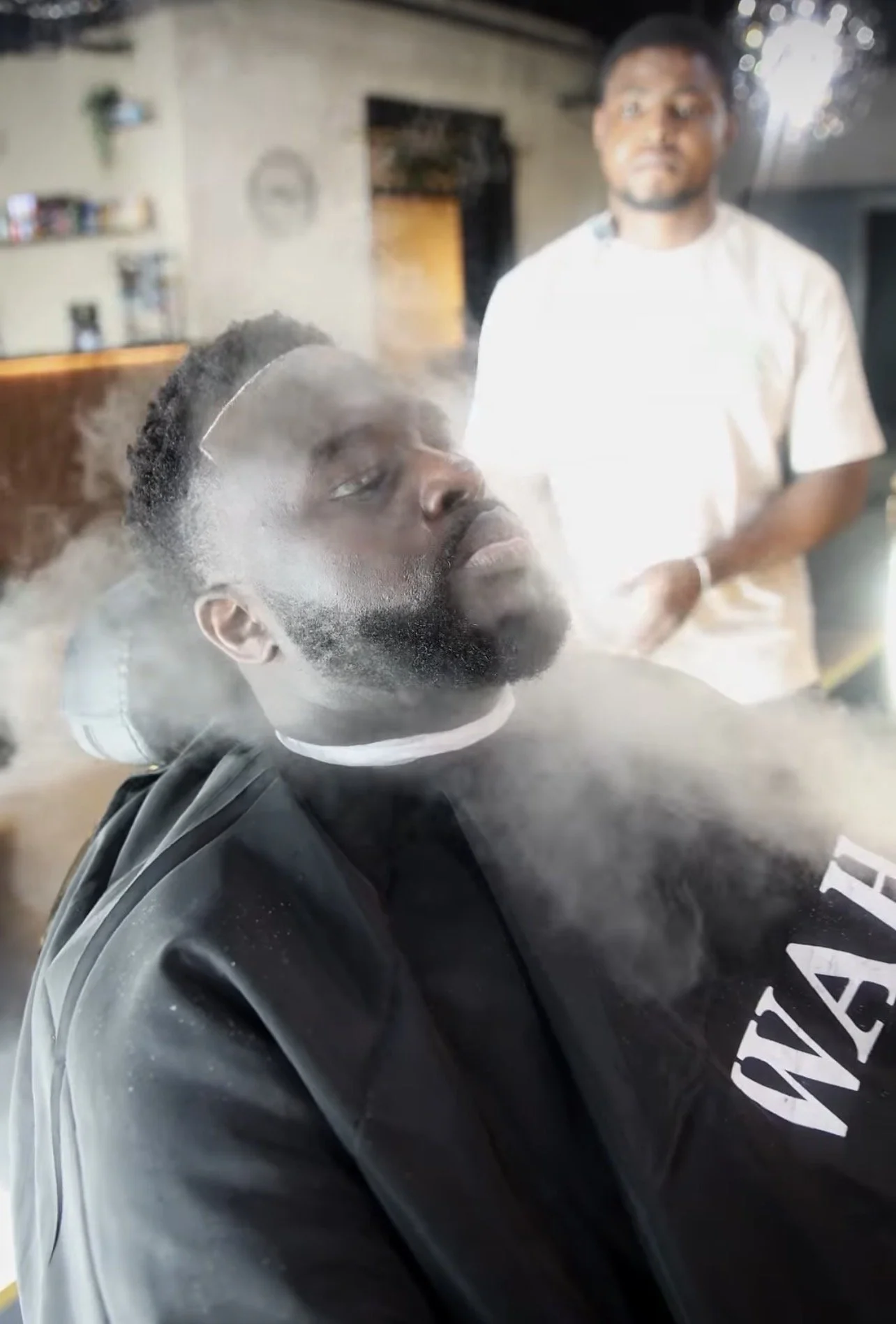 A man with a beard and a headband getting a haircut with steam or fog around his head, while another person stands in the background of the salon.
