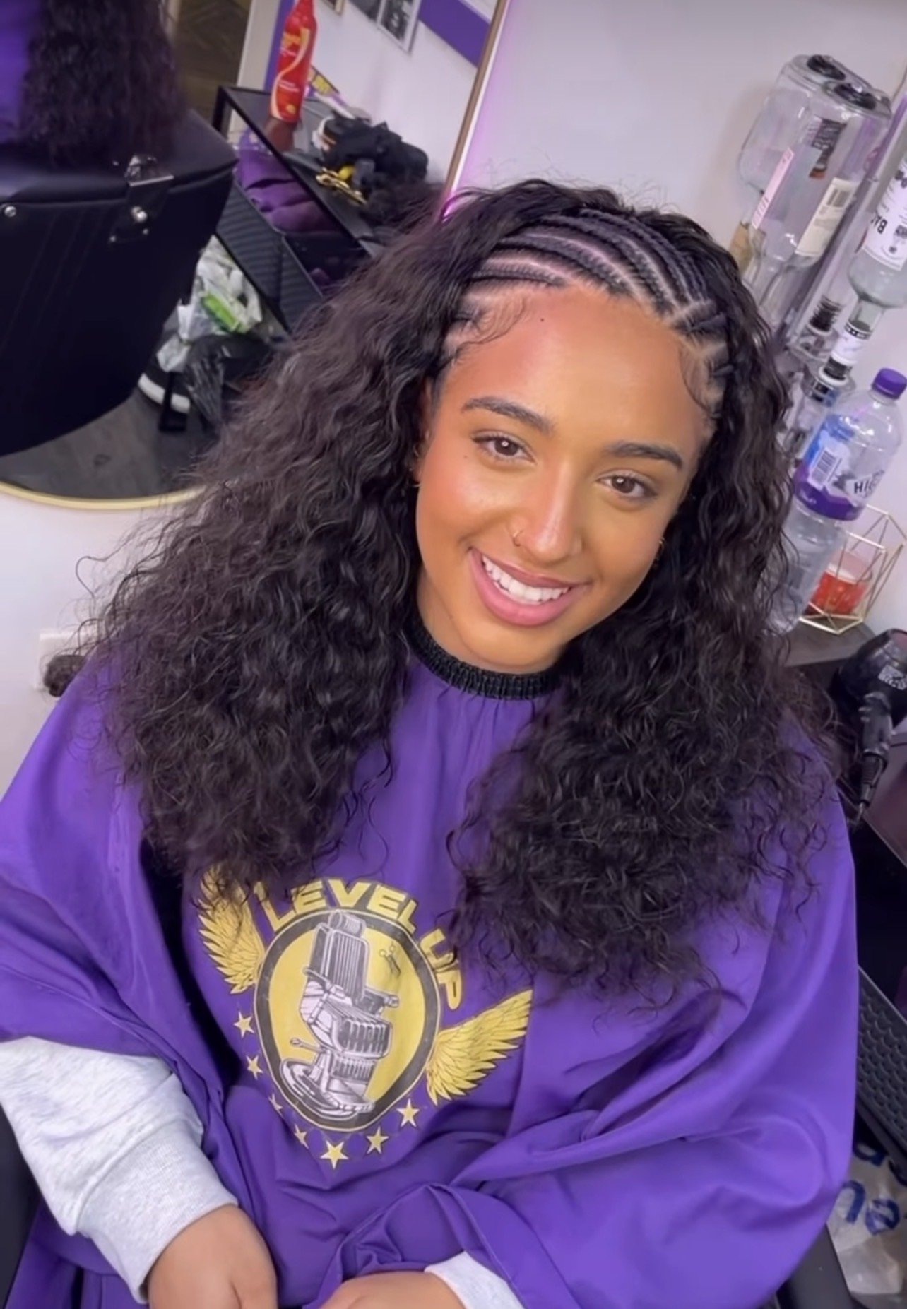 Woman with curly hair and a nose piercing smiling, wearing a purple sweatshirt with a logo, sitting in a salon chair. In the background, a mirror, water bottles, and a hairdresser's station are visible.