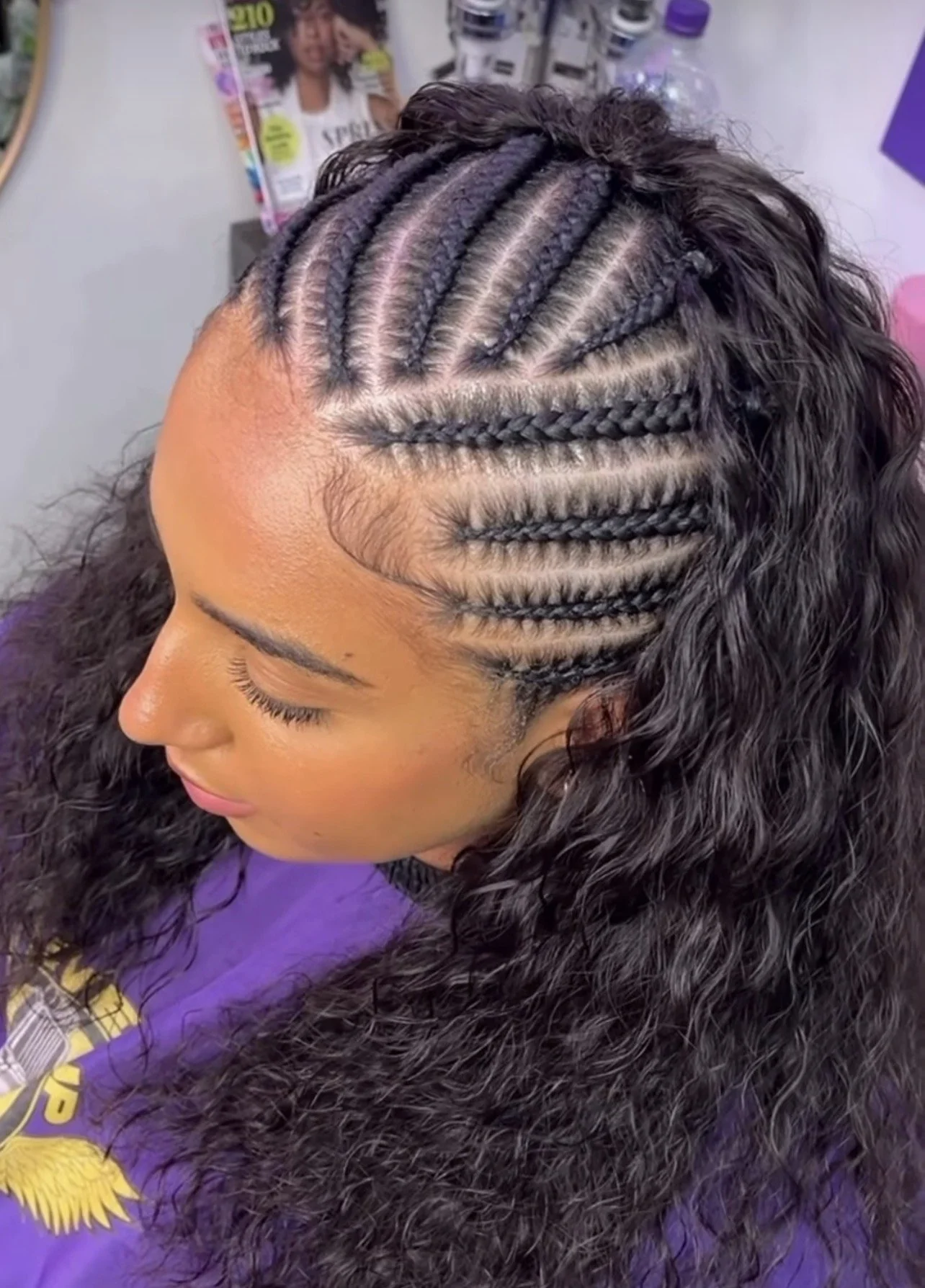 A woman with styled cornrow braids on her scalp, showing intricate parting lines. She has long, curly hair and makeup, and is wearing a purple shirt with a yellow design.