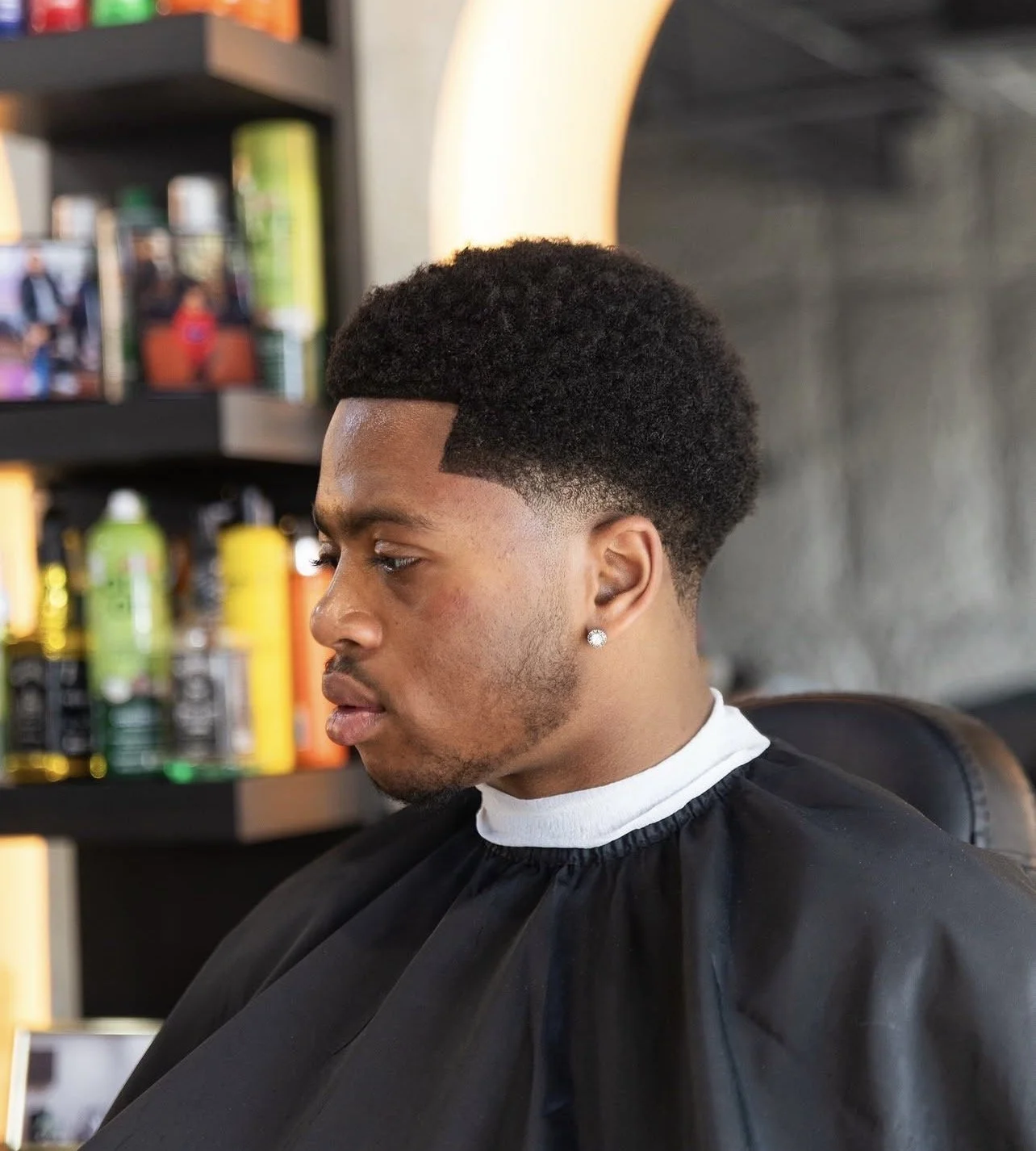 Young man with a fresh fade haircut sitting in a barbershop, wearing a black cape, with hair products on shelves in the background.