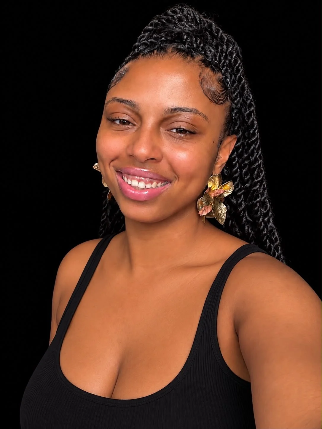 A woman with dark, curly hair styled in twists, wearing large floral earrings and a black tank top, smiling against a black background.