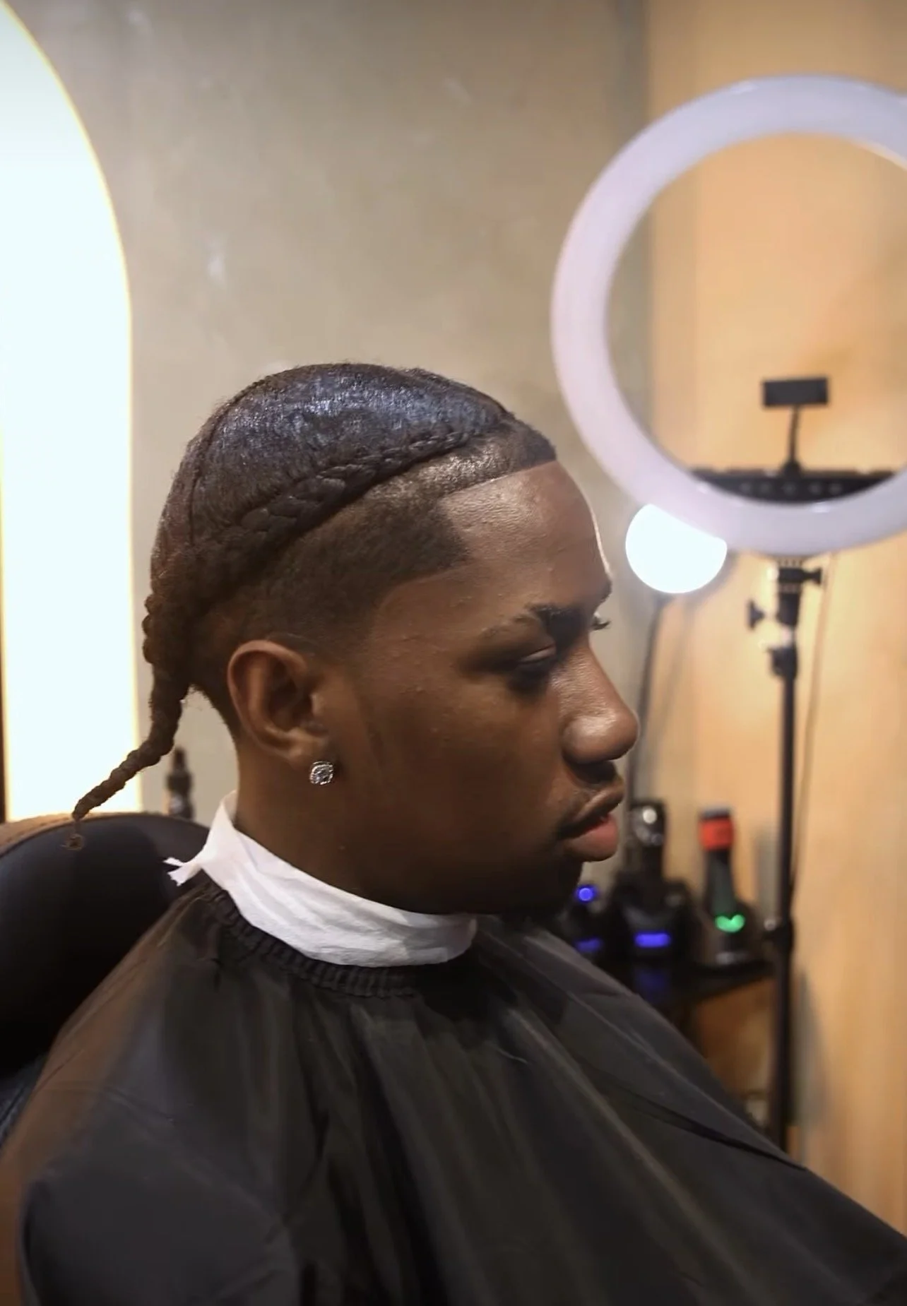 A man with neatly styled hair and a braid on top, wearing earrings and a white shirt, sits in a barber's chair in front of a mirror, getting a haircut or styling.