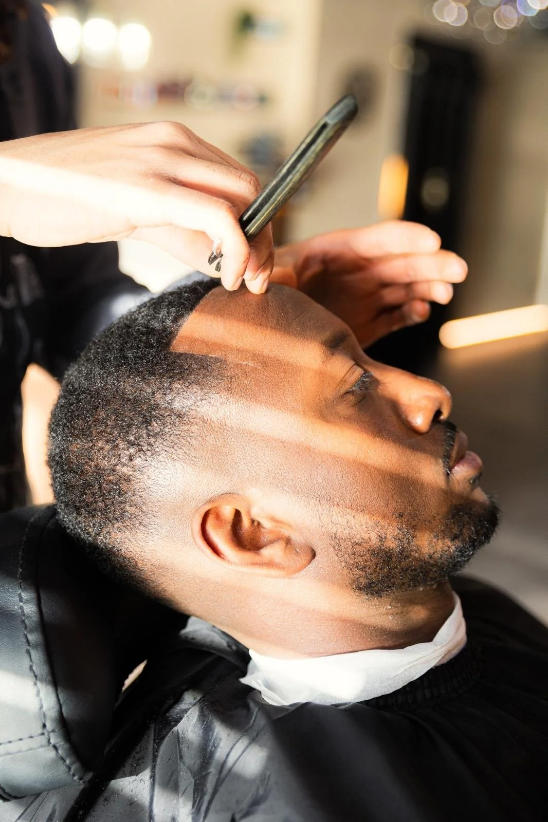 Man receiving a haircut, with lines shaved into his hair and a barber using a clipper, in a barbershop.