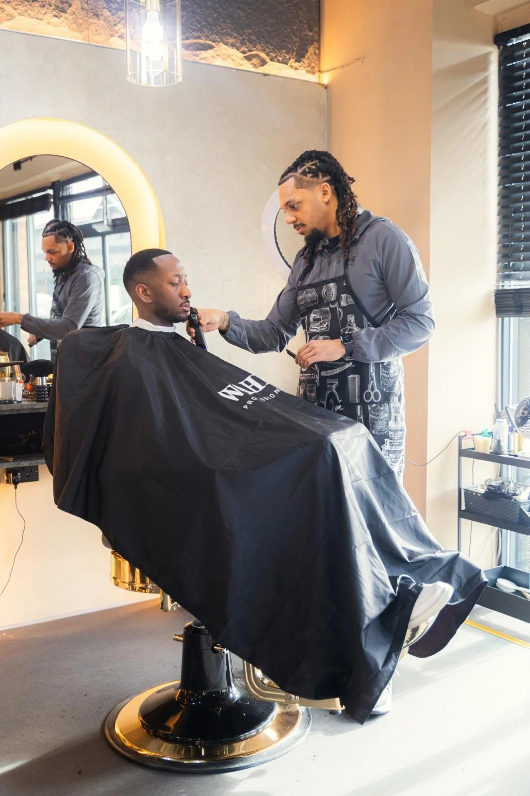 A man getting a haircut at a barbershop, seated in a barber chair with a black cape. A barber stands beside him, holding a straight razor, preparing to trim the man's beard. The shop interior includes a mirror, barber tools, and another man working at a station in the background.