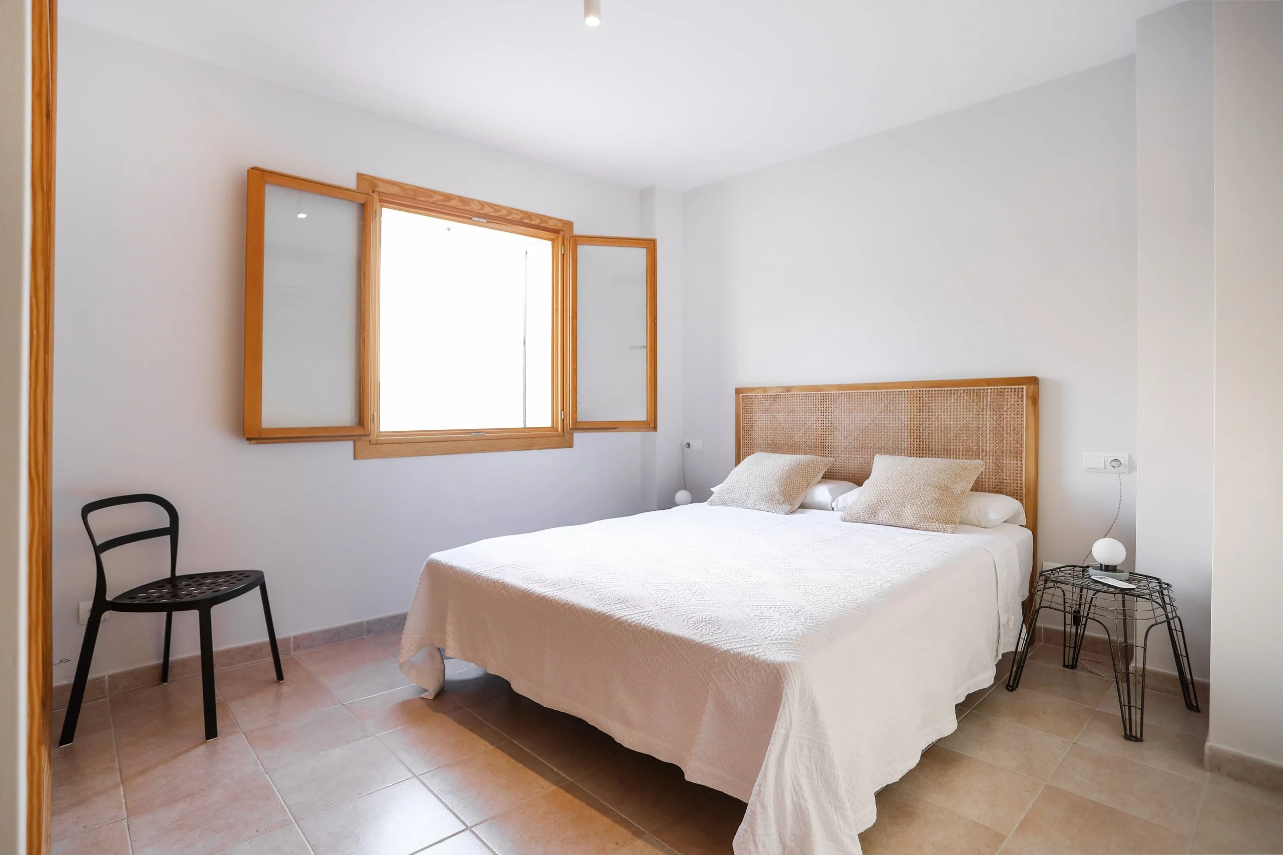 Habitación con cama blanca, cabecera de mimbre, dos almohadas claras, ventana de madera abierta, silla negra, lámpara de mesa en mesa de noche de metal, paredes blancas, suelo de baldosas beige.