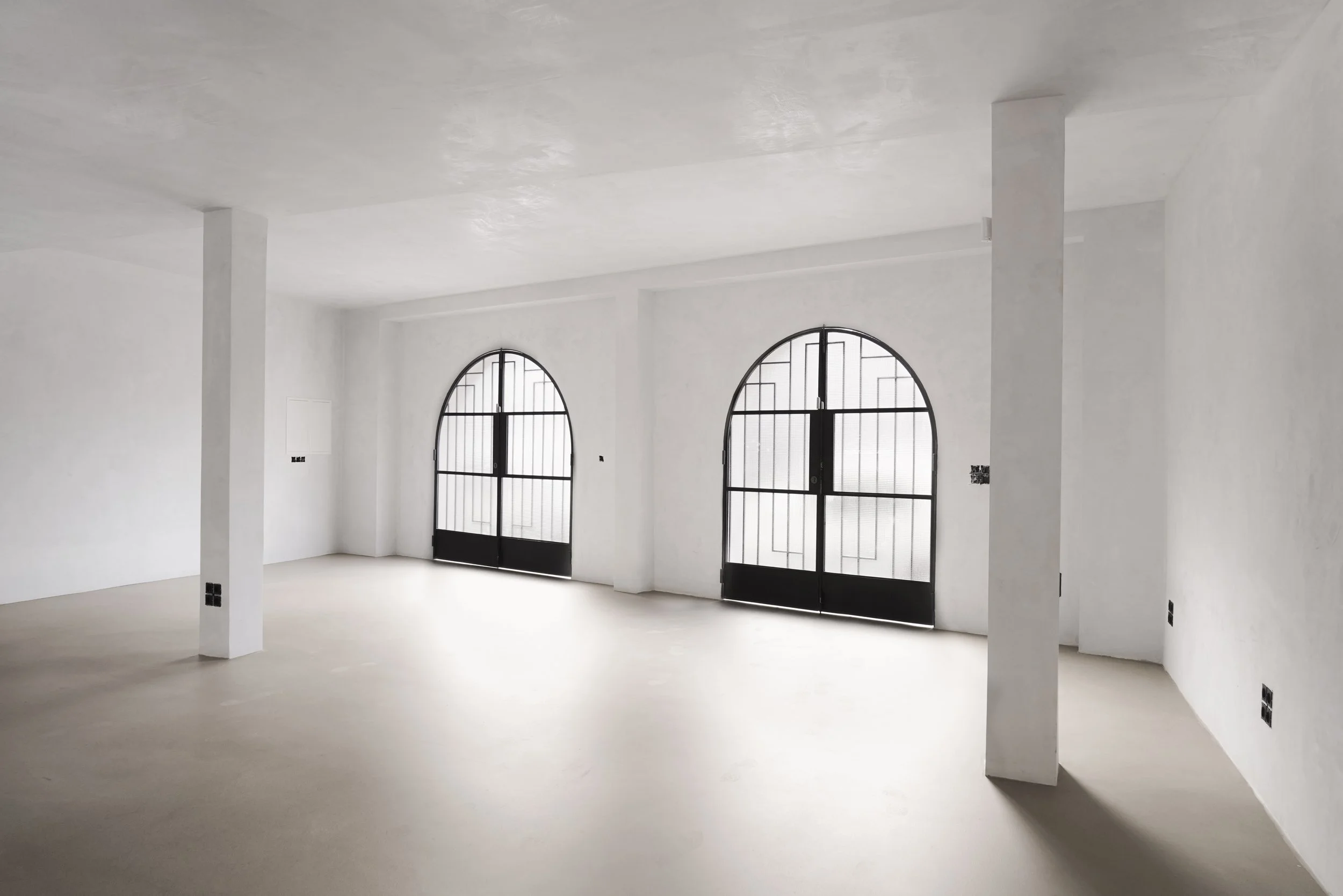 Interior de un espacio vacío con paredes y techo blancos, puertas metálicas negras con arcos y rejas, y ventanas con rejas de diseño geométrico.