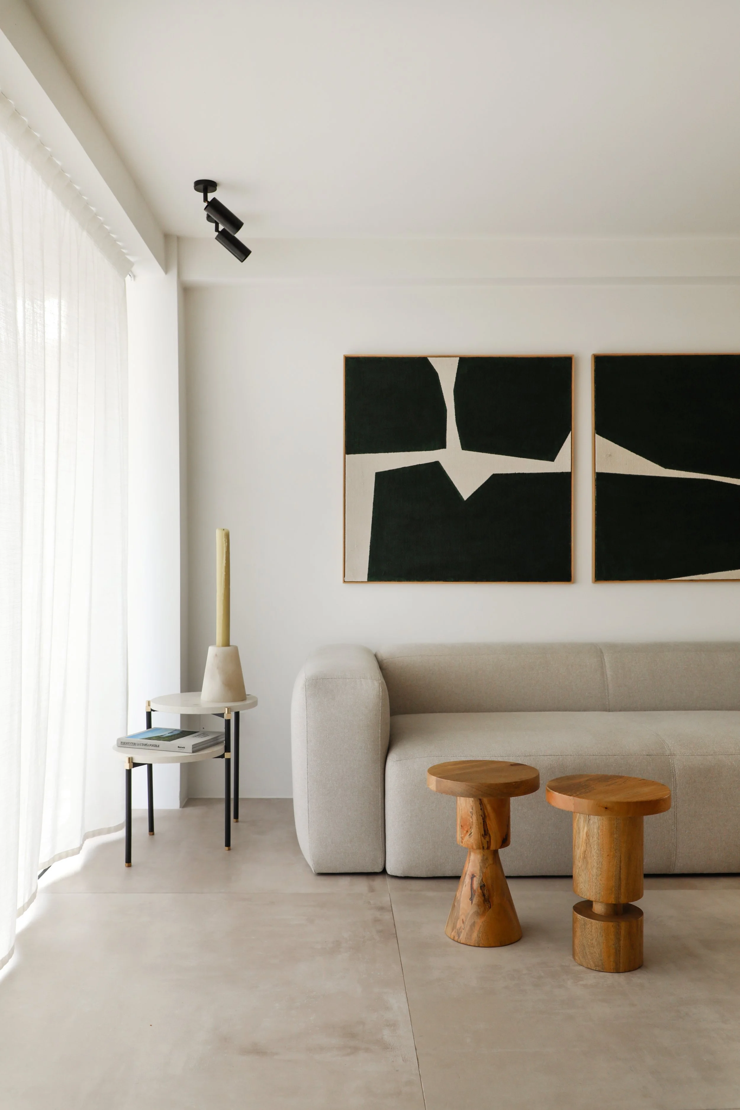 Sala de estar con pared blanca, dos cuadros abstractos en tonos negro y blanco, sofá beige, mesa de madera, lámpara de pie y mesa auxiliar con libros.