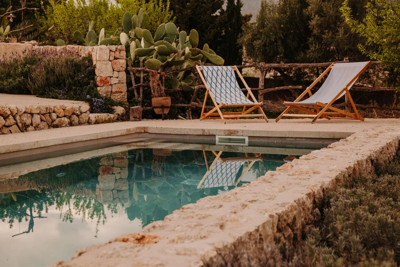 Two wooden deck chairs with fabric seats sit next to a small swimming pool, surrounded by stone and lush green plants.