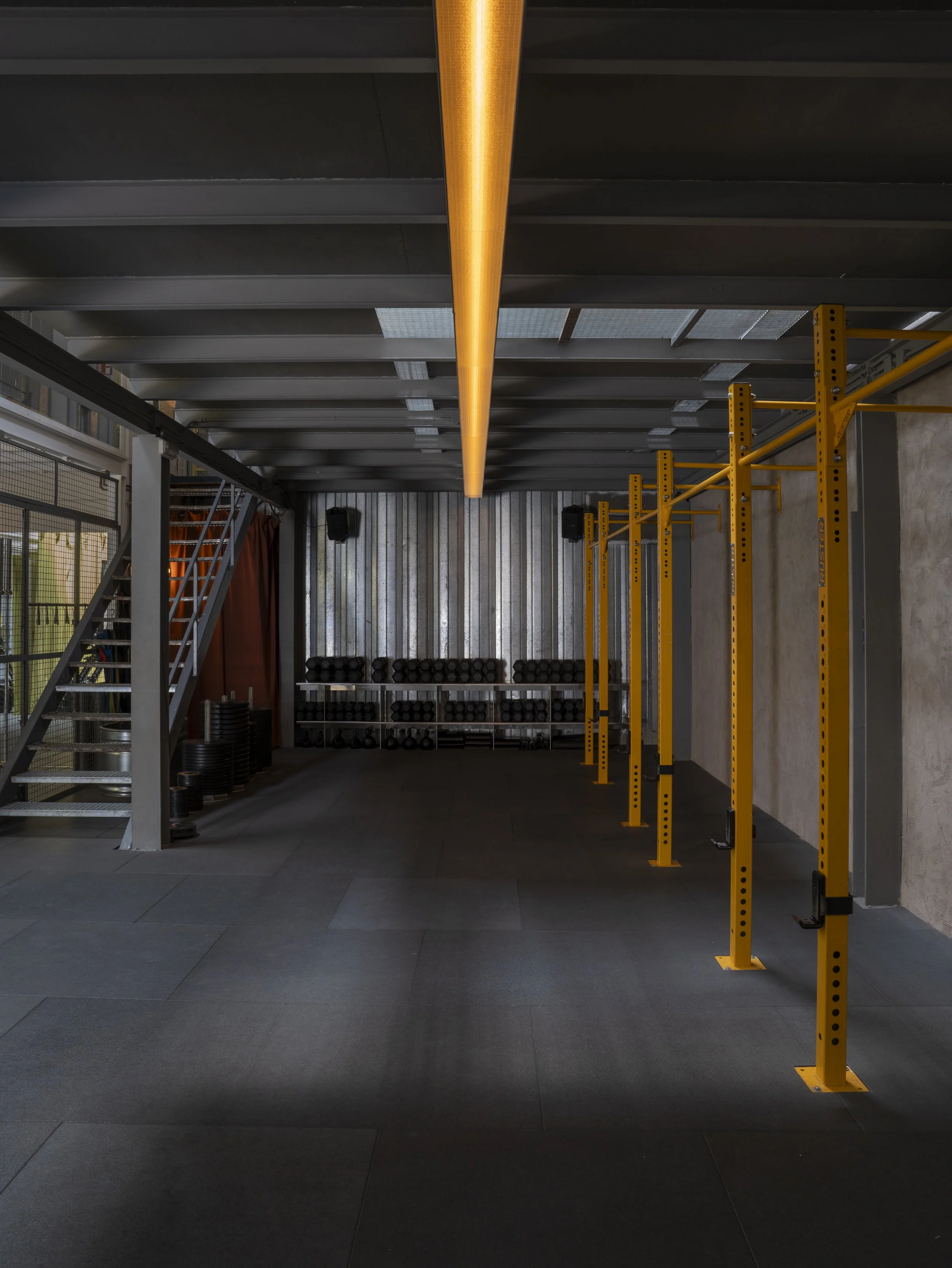 Espacio de gimnasio con estructura de estilo industrial, barras de peso, estantería con pesas, suelo de baldosas oscuras y paredes de metal, con una larga luz decorativa en el techo.