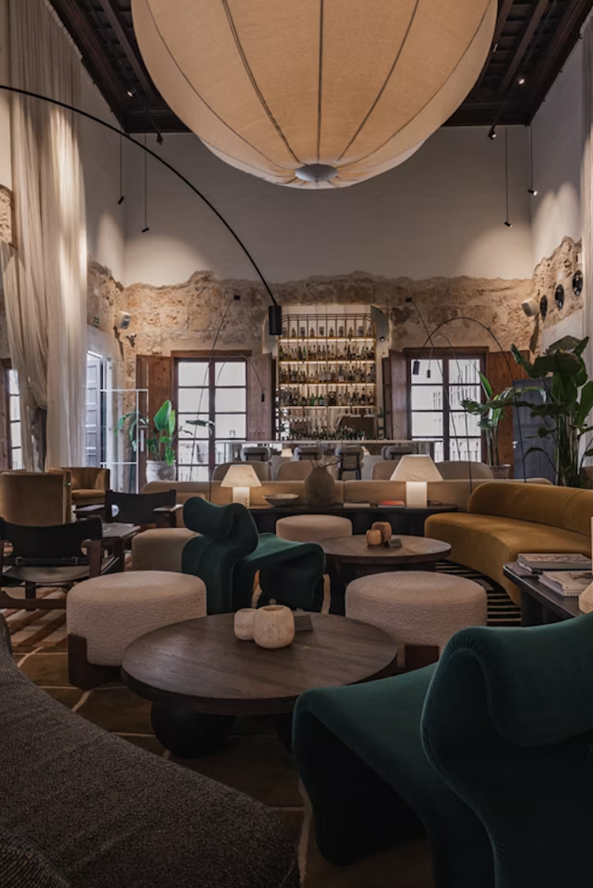 Interior de un salón elegante con muebles cómodos, lámparas y plantas, paredes de piedra y ventanas grandes con cortinas claras, decorado con estilo moderno y acogedor.