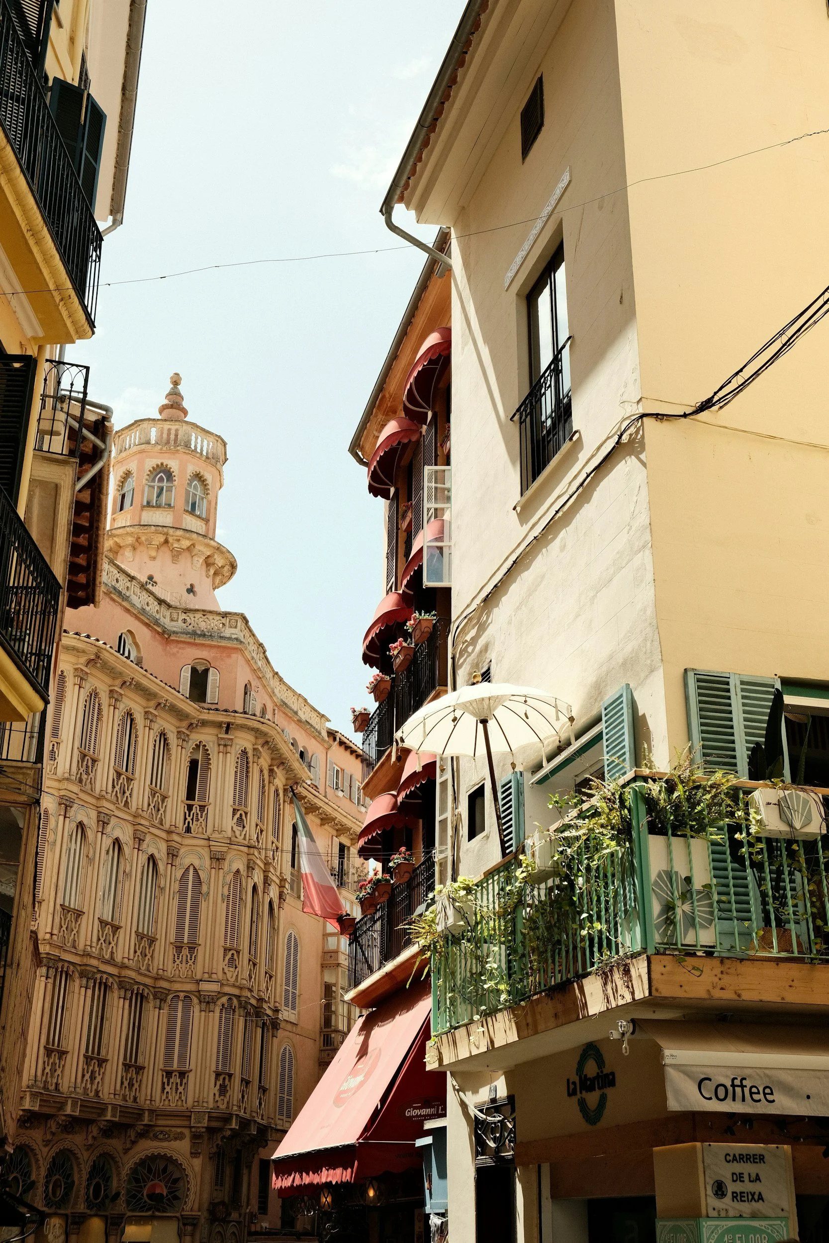 Vista de una calle en una ciudad con edificios antiguos y modernos, tiendas, balcones con flores, y un cielo despejado.