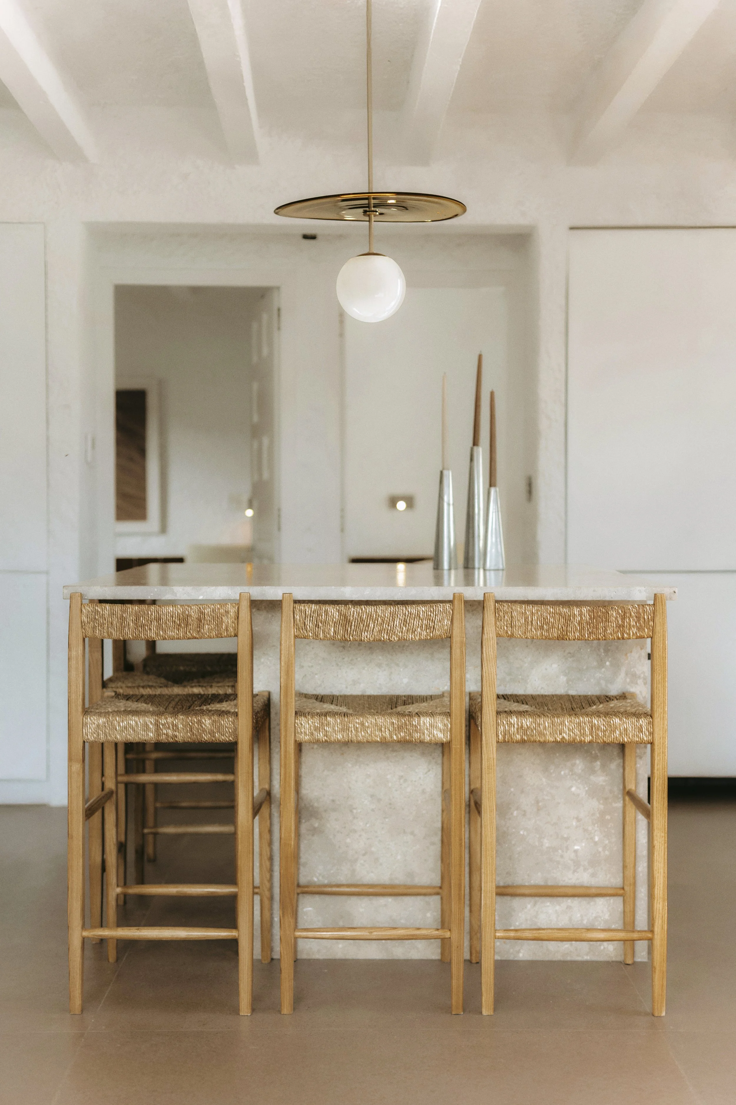 Mesas de comedor en un espacio interior minimalista con sillas de madera y asiento de cuerda, decorado con jarrones altos en tonos neutros y una lámpara moderna en el techo.