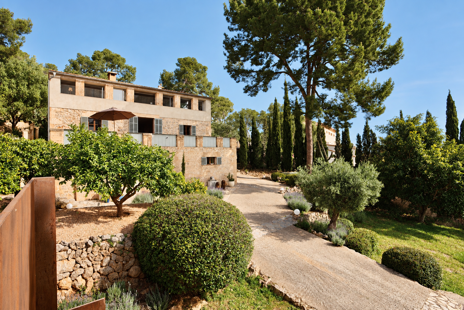 A hillside residence with stone and stucco exterior surrounded by lush green trees and garden plants, under a clear blue sky.