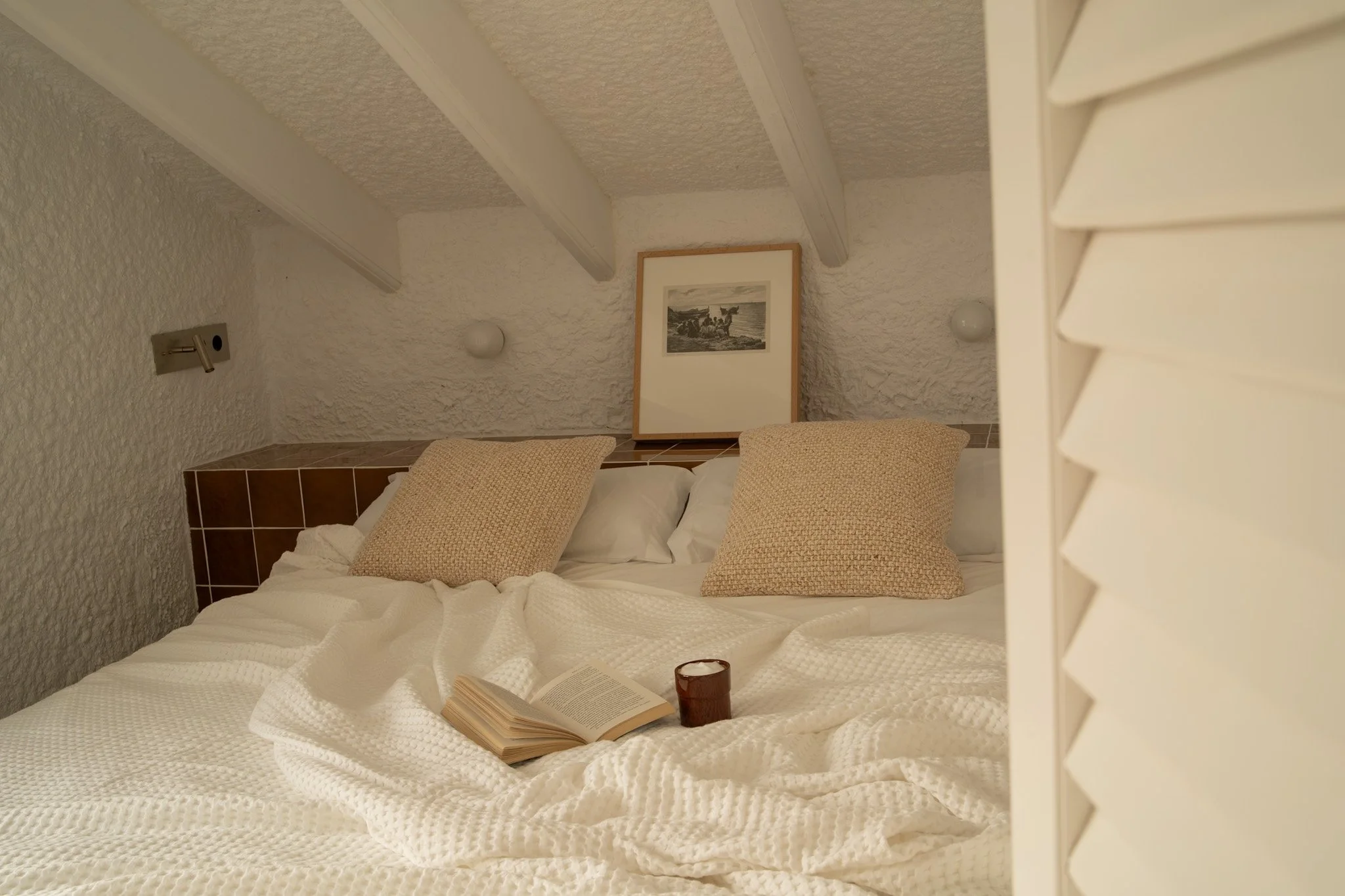 Cama con almohadas y libro abierto, taza de color oscuro, en habitación con paredes de textura irregular y un cuadro en la pared.