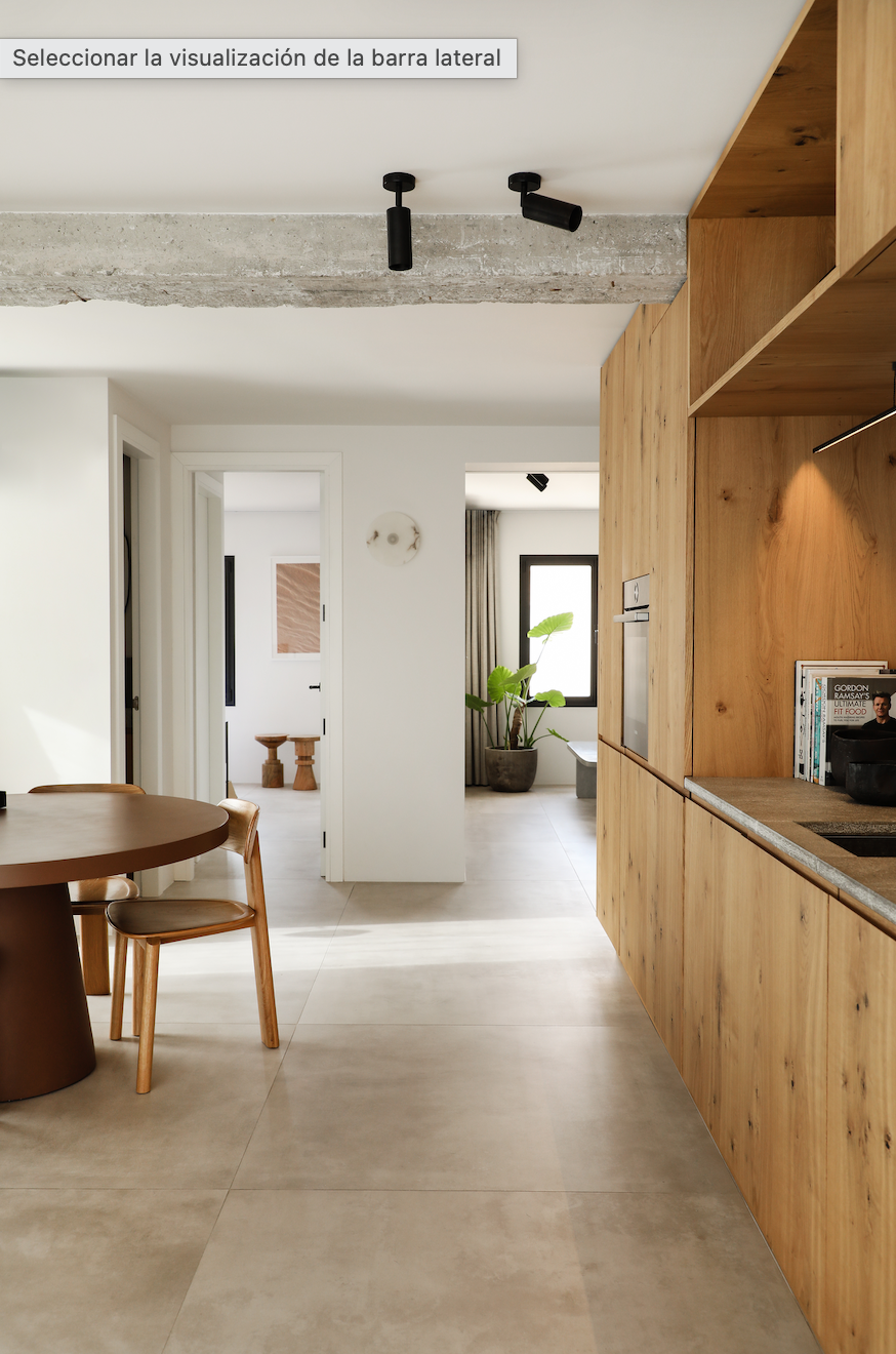 Interiores modernos de una casa con paredes blancas, suelo de baldosas beige, mueble de cocina de madera, mesa redonda de madera y sillas del mismo material, plantas en decoración, ventanas grandes con cortinas, y techos con iluminación negra y estructura de concreto.