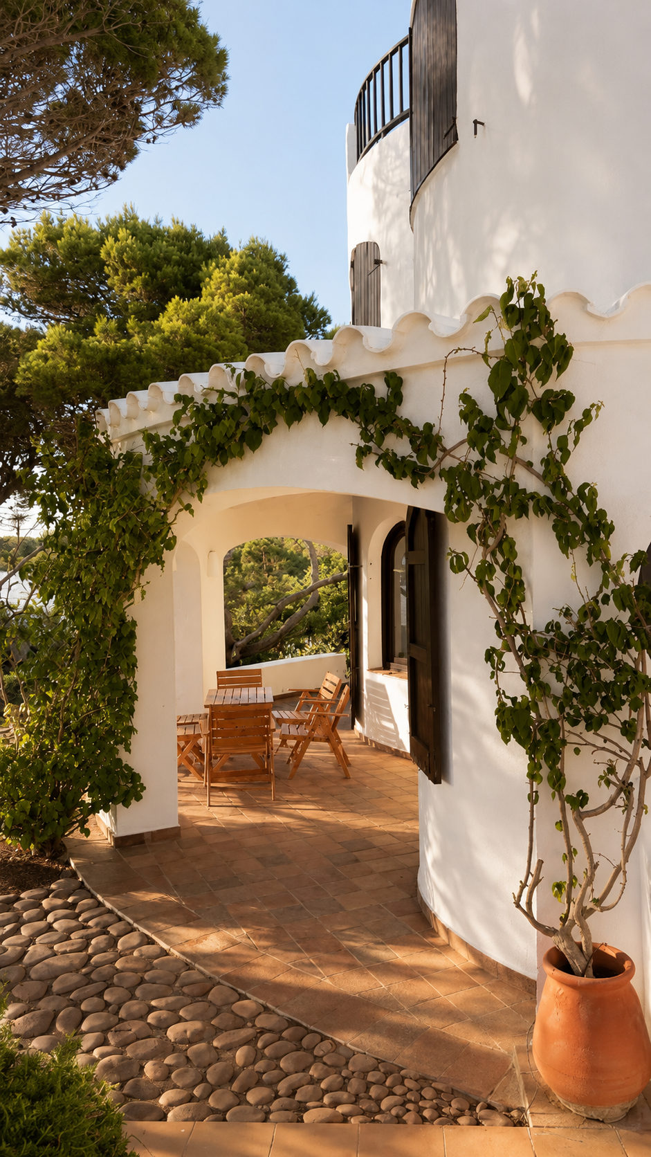 Casa blanca con terraza decorada con plantas y mobiliario de madera, rodeada de árboles y jardines.