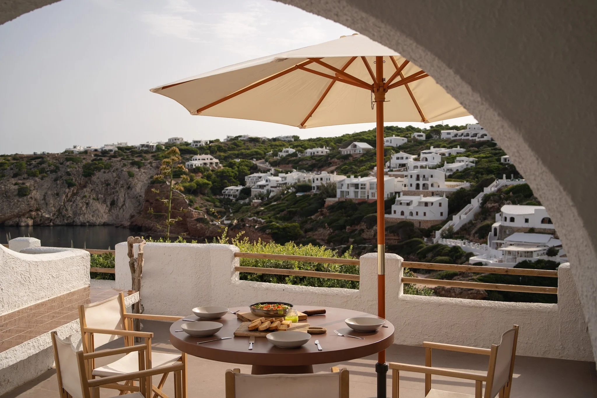 Vista de una mesa redonda en una terraza con platos, un cesto con pan, y utensilios de comida. En el fondo, vista de casas blancas en una colina y un paraguas beige.