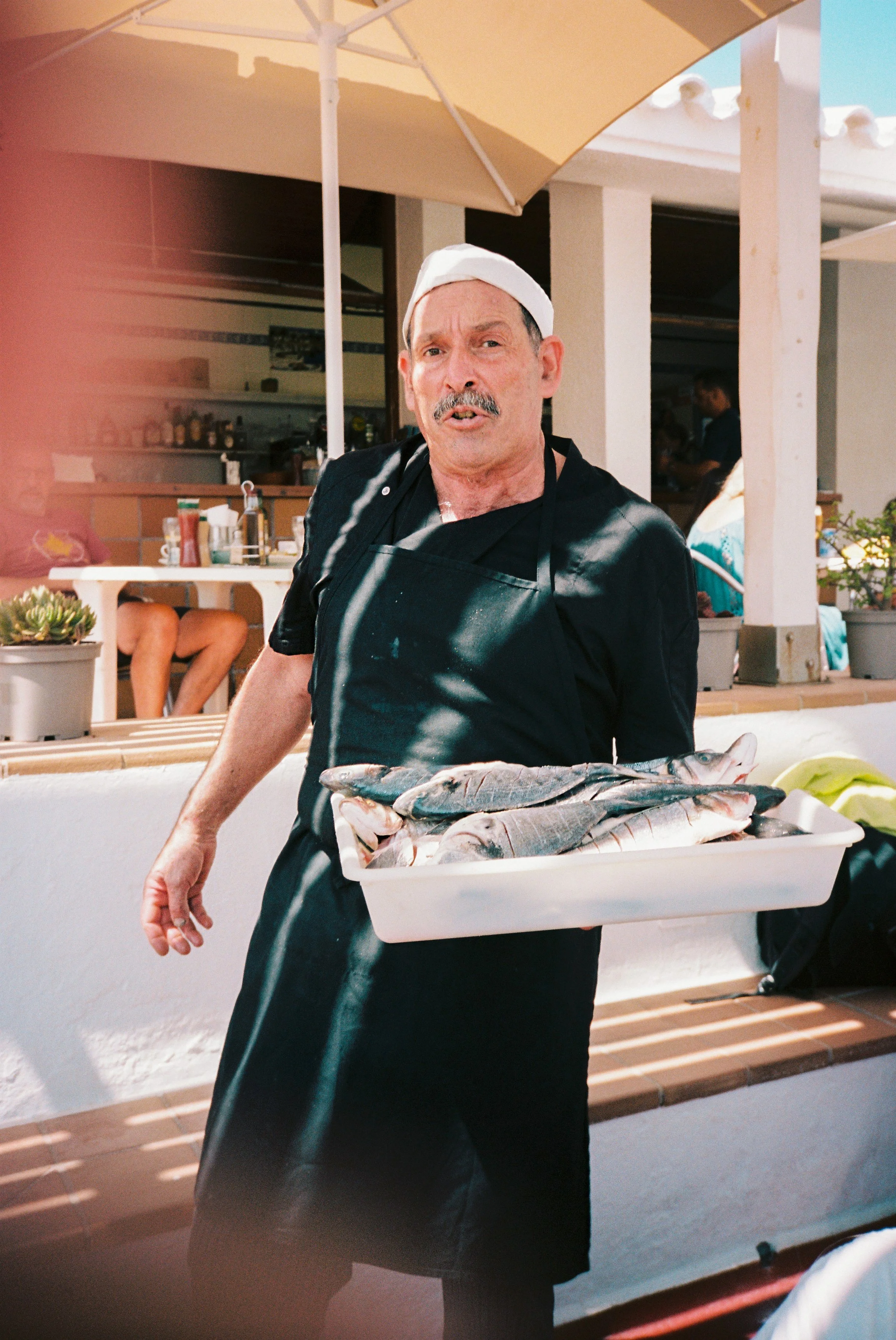 Hombre sirviendo pescado en un restaurante o feria al aire libre bajo una sombrilla de color beige, con una barbacoa y plantas decorativas en el fondo.