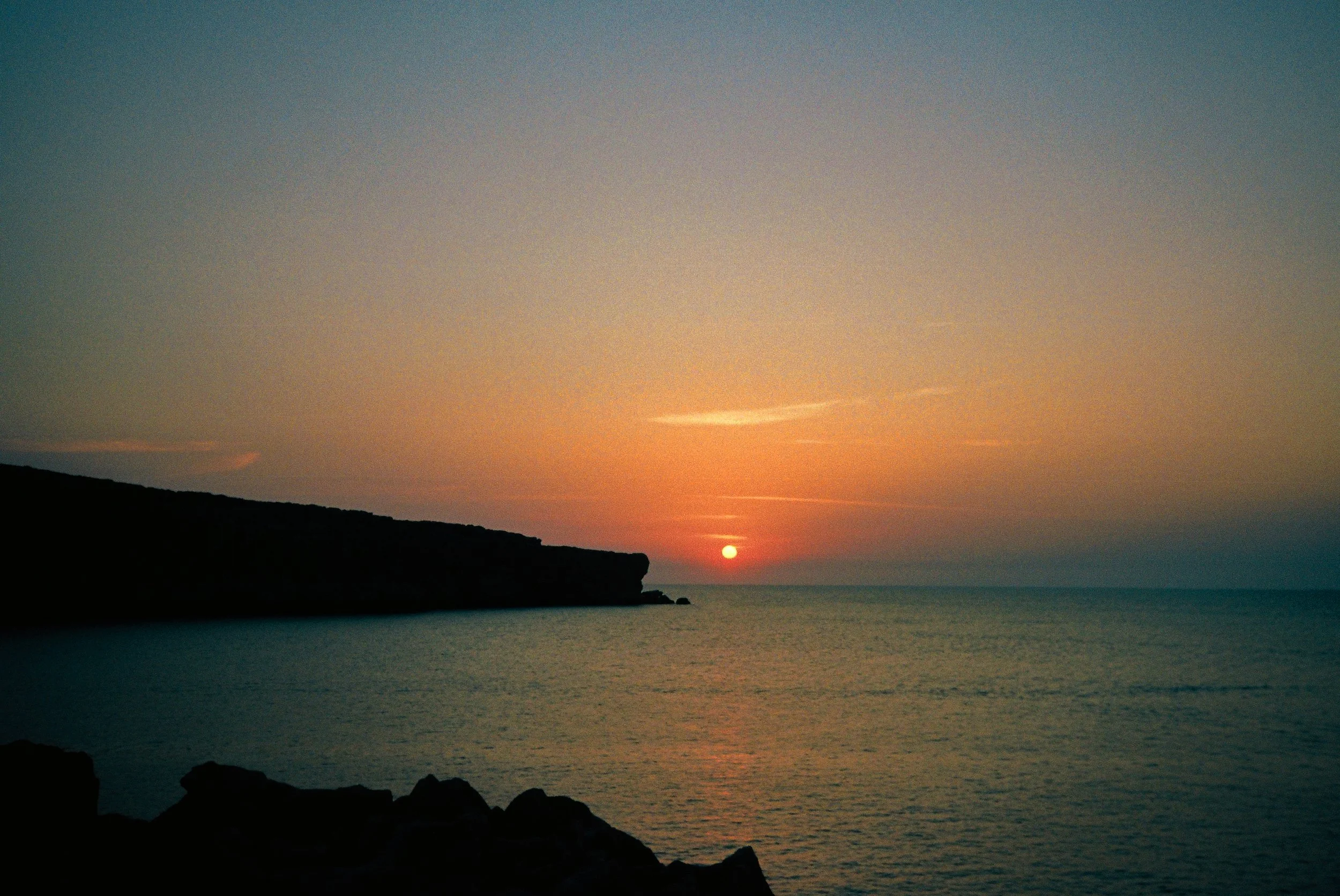 Atardecer en la playa con el sol bajando sobre el horizonte, nubes dispersas y una silueta de acantilados en la orilla del mar.