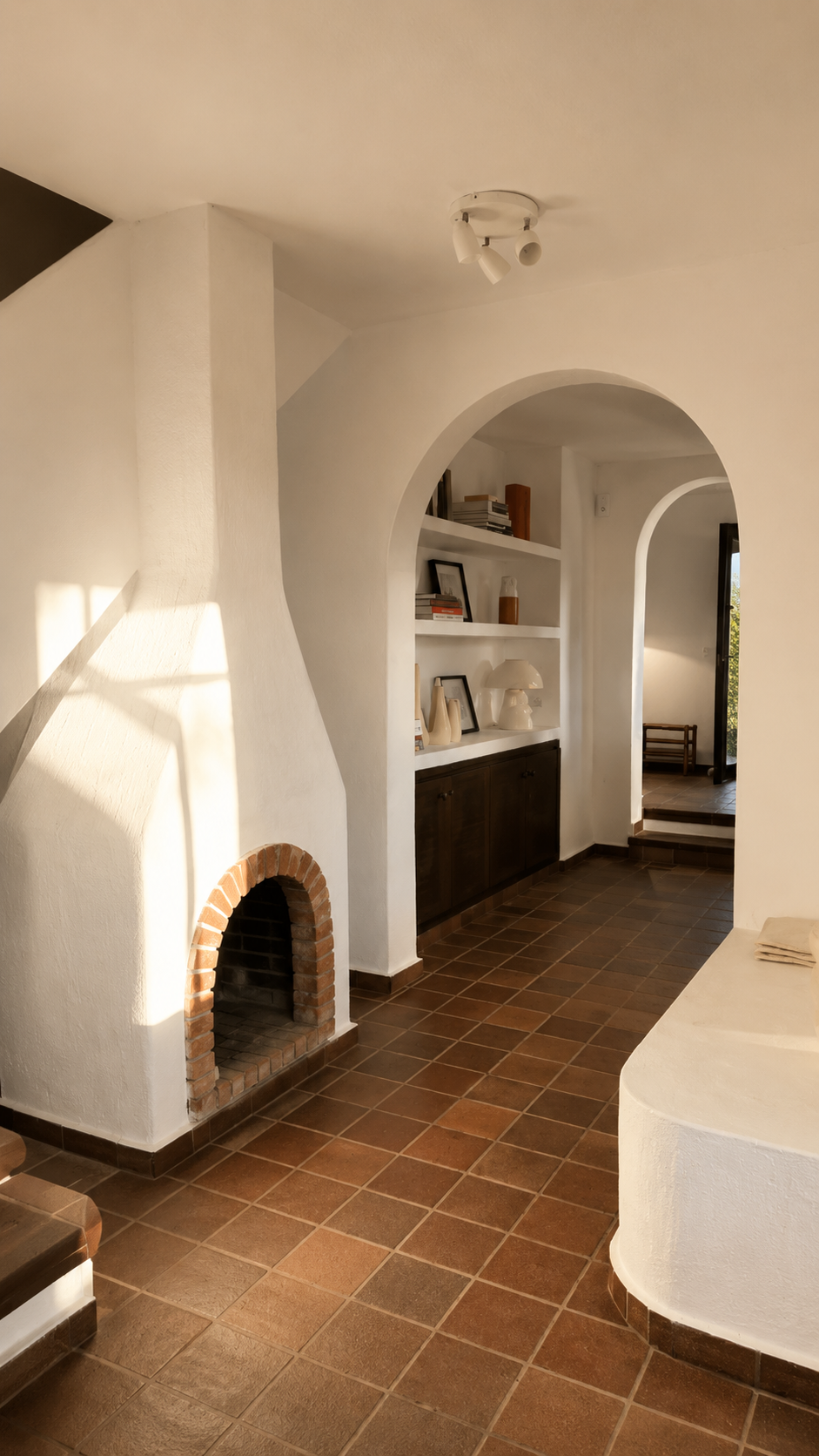 Vista interior de una casa con paredes blancas, suelo de baldosas y decoraciones en estantes blancos, incluyendo libros, fotos y lámparas, con espacio para una chimenea de ladrillo y un arco interior que conduce a otra habitación.