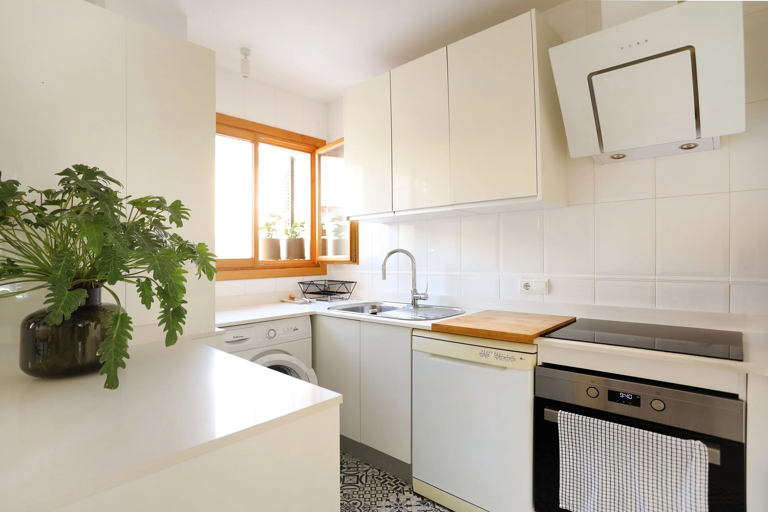 Cocina moderna con armarios blancos, ventanas de madera, electrodomésticos, y planta en un jarrón oscuro en una superficie blanca.