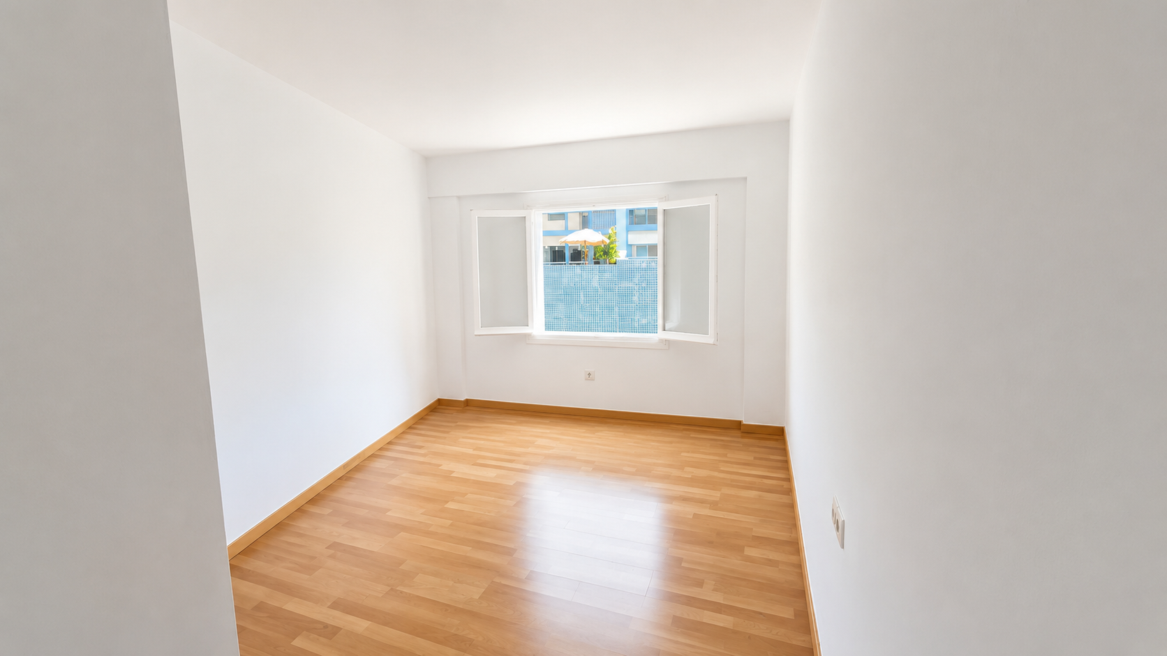 Habitación vacía con paredes blancas, piso de madera y una ventana abierta que muestra un balcón con sombrilla y plantas.