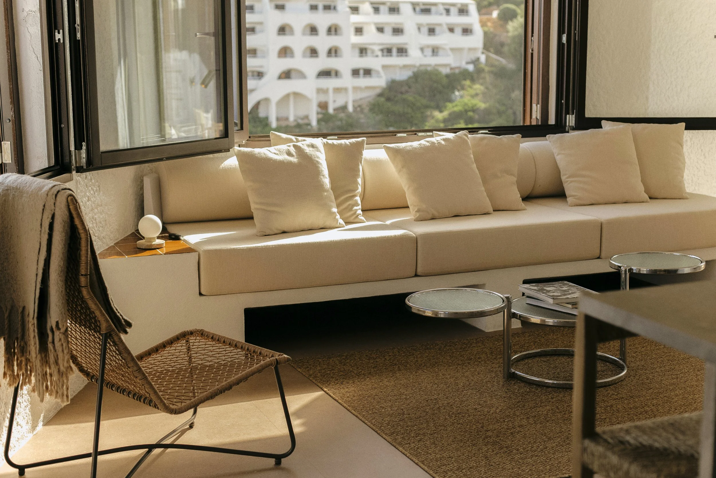 Sala de estar con sillón blanco, múltiples almohadas, mesa de centro y ventanas abiertas con vista a edificios y árboles.