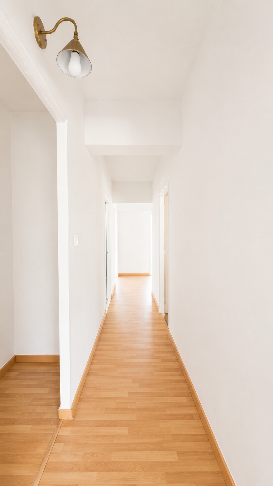 Pasillo interior con paredes blancas, suelo de madera clara, una lámpara en la esquina superior izquierda, y varias puertas a los lados.