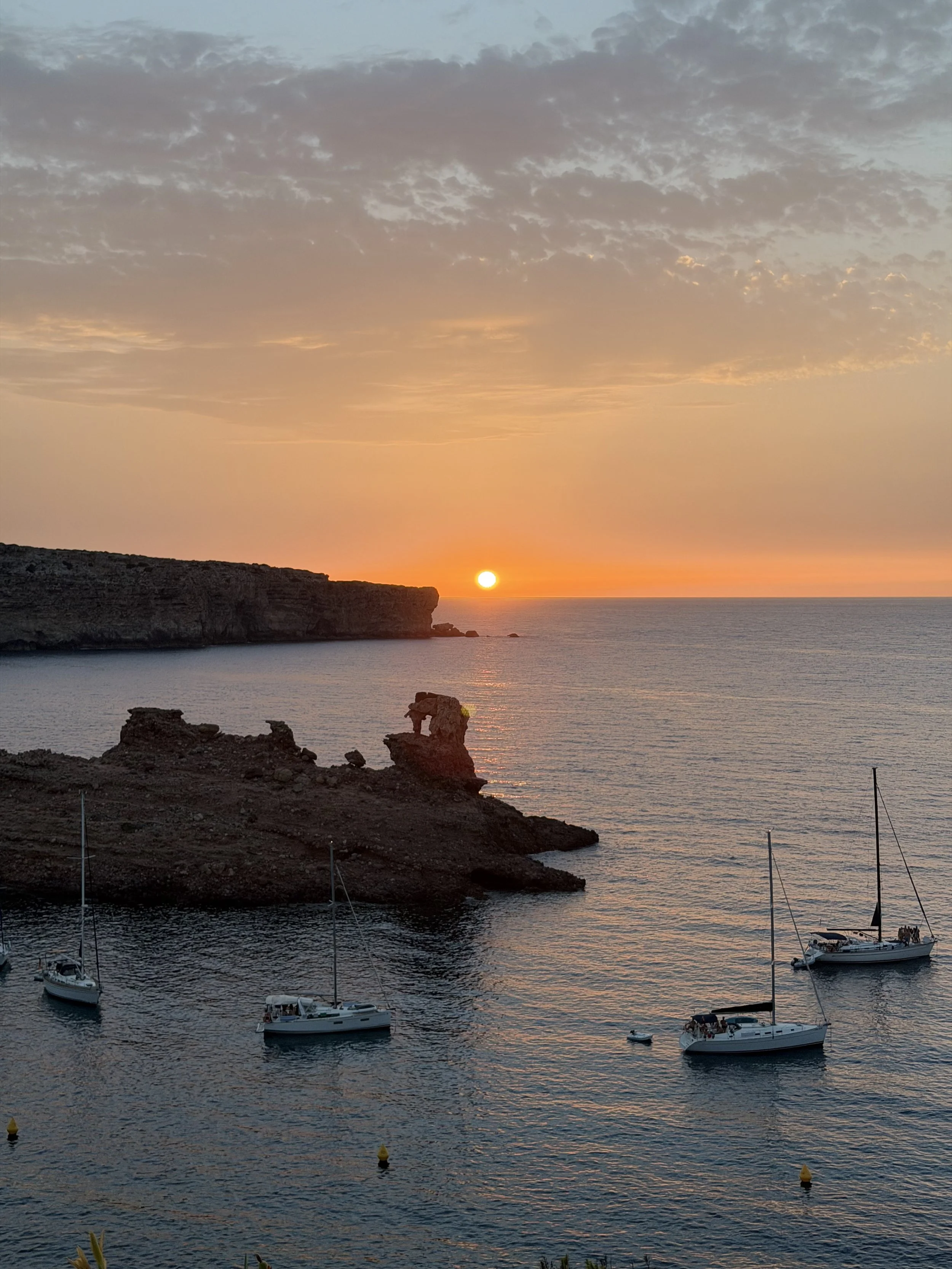 Vista marítima al atardecer con varias embarcaciones fondeadas en el agua y un peñón en la costa, con el sol poniéndose en el horizonte y cielo con nubes dispersas.
