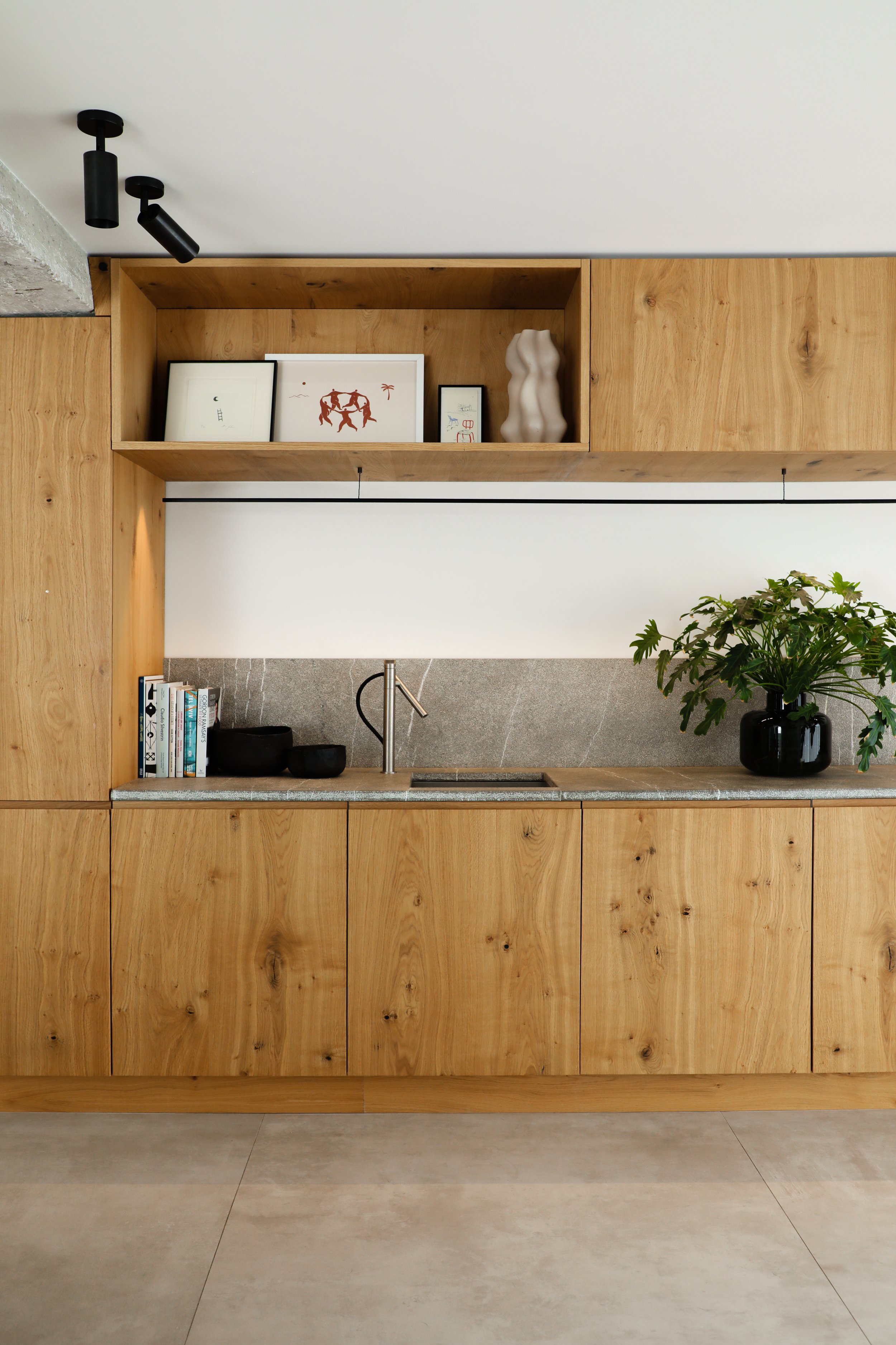Vista de una cocina moderna con gabinetes de madera, una encimera de piedra, y decoración minimalista, incluyendo libros, plantas y cuadros en la estantería.