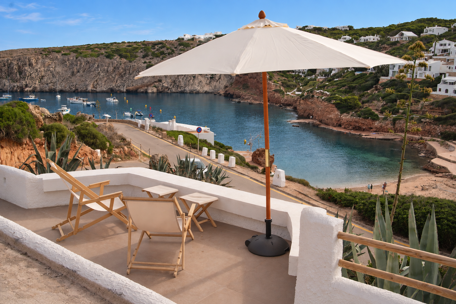 A balcony overlooking a bay with boats, surrounded by rocky cliffs, with a large white beach and white buildings on the hillside in the background. There is a large umbrella and two wooden chairs on the balcony.